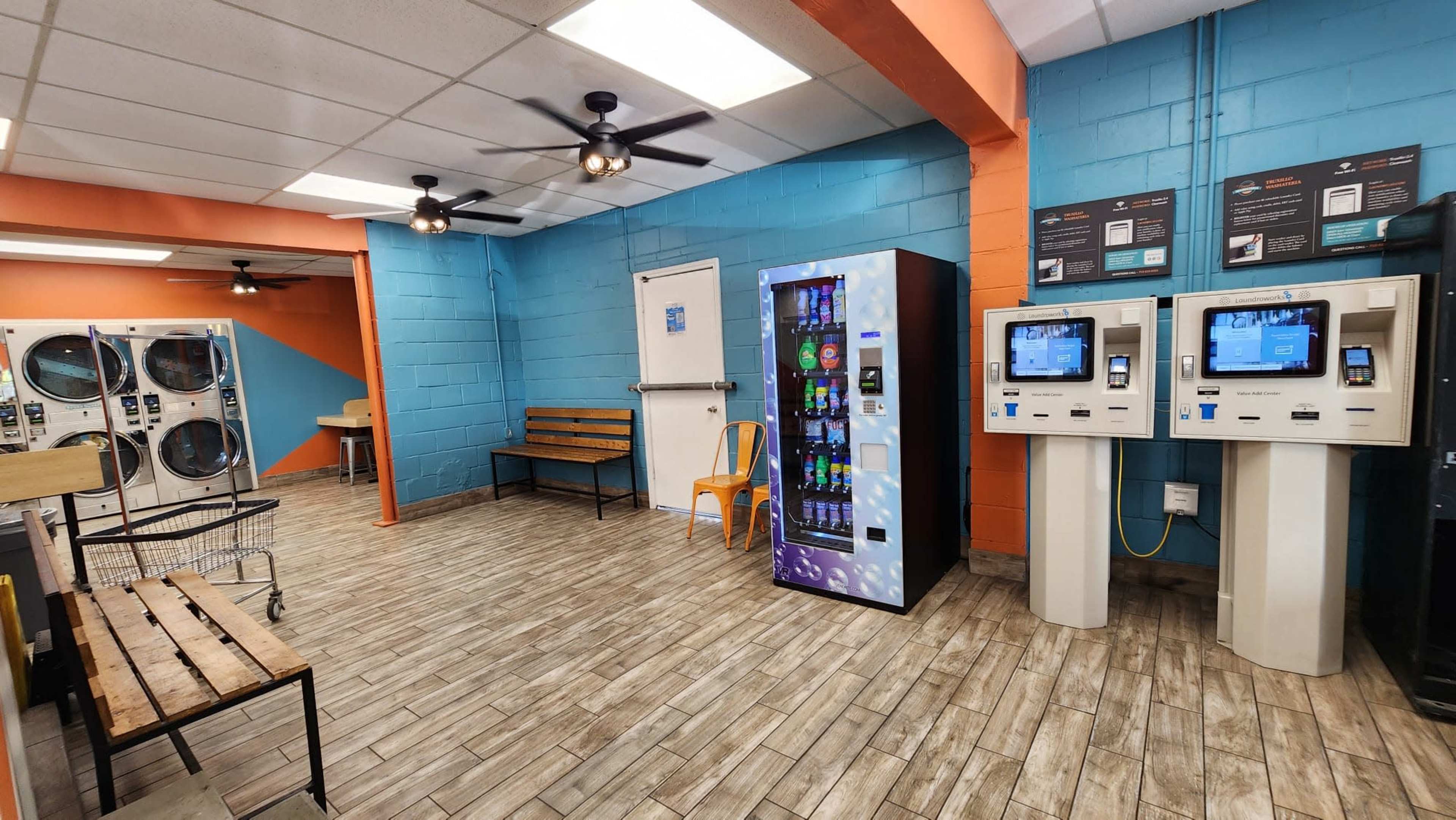 An empty laundromat with a vending machine, two self-service kiosks, and laundry machines along the walls.