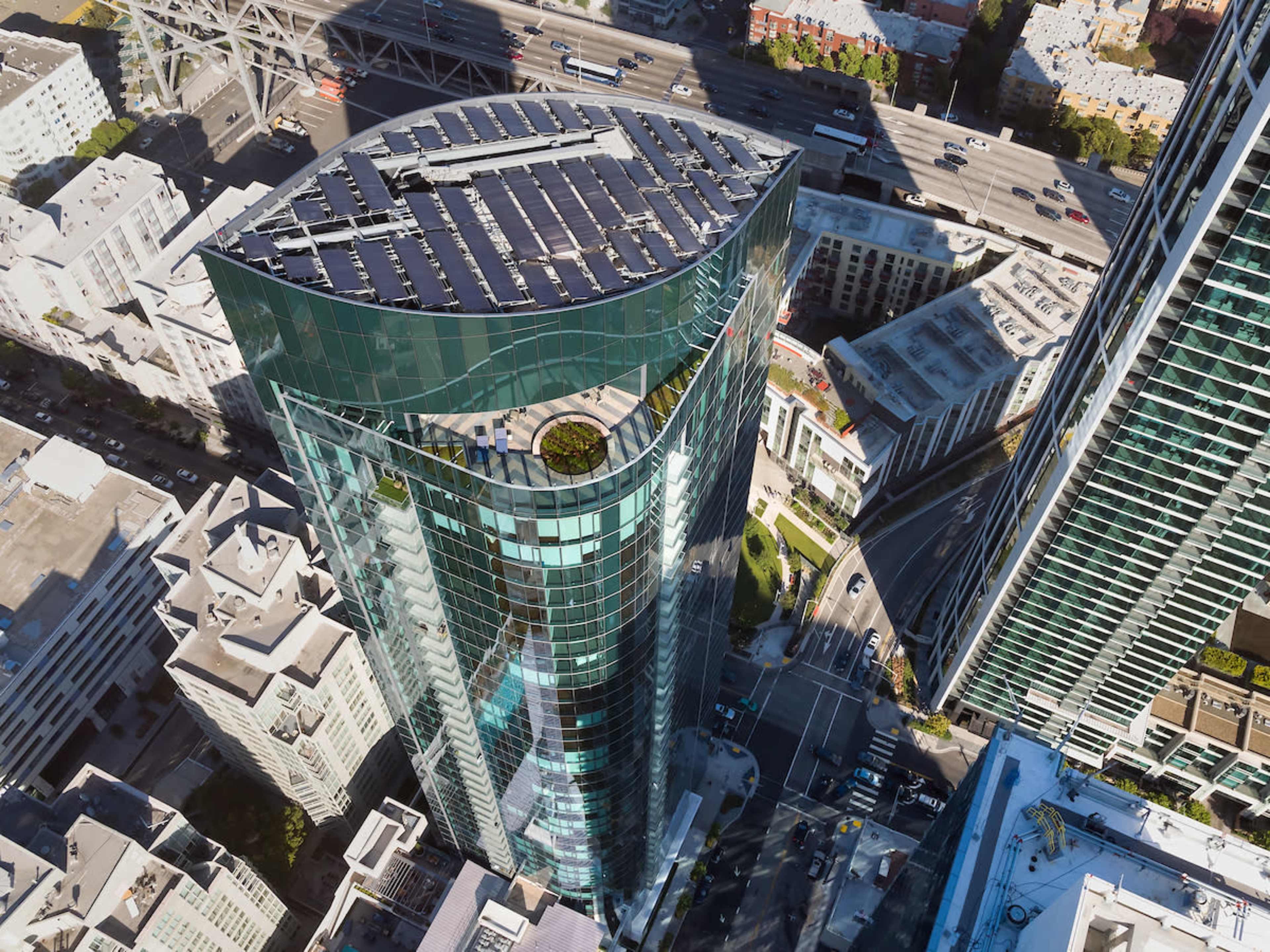 A modern high-rise building with a curved glass facade and a green rooftop features is situated among city streets and other structures.