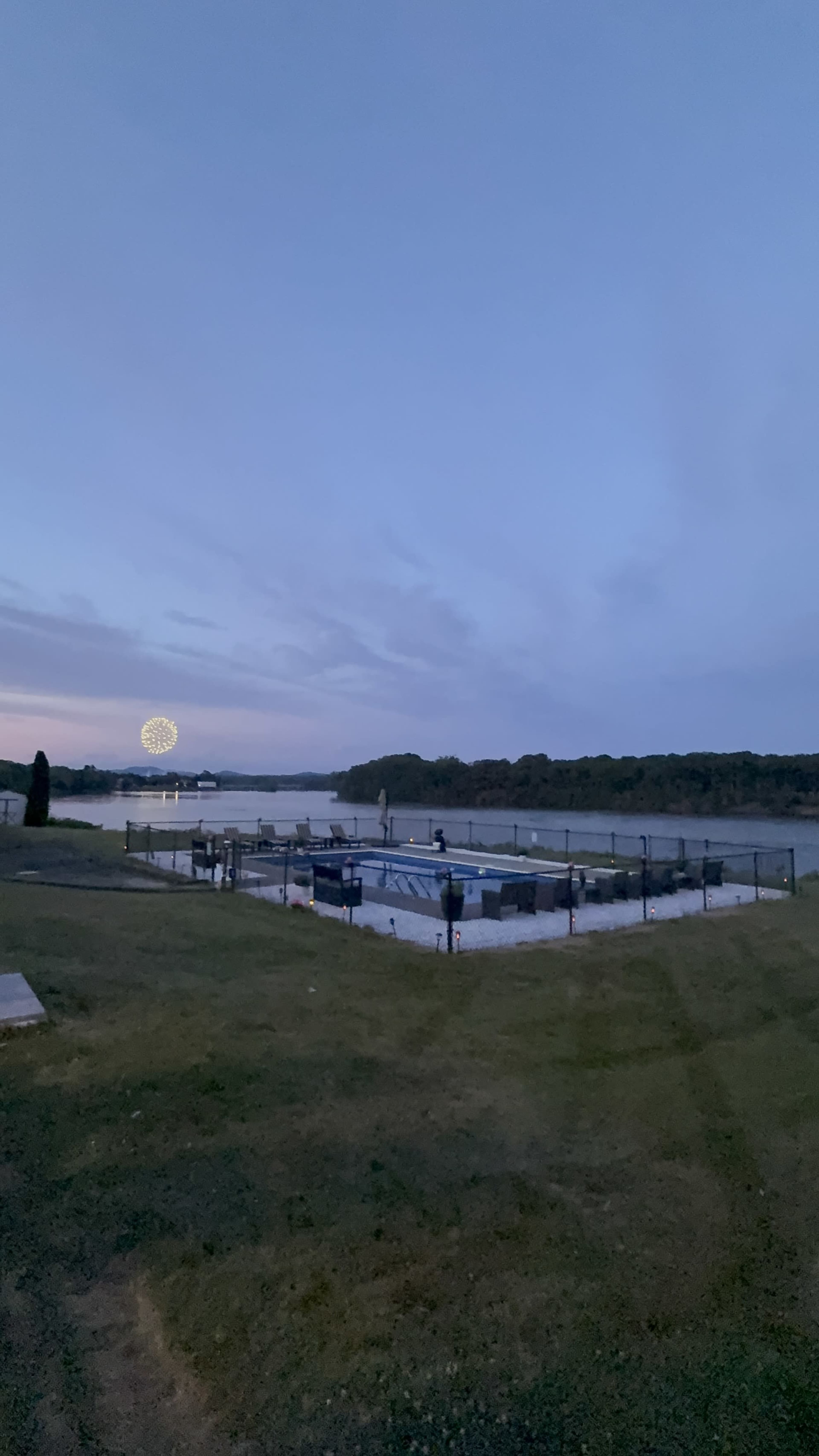 The image shows a view of a calm waterfront area with a pool and dock, under a twilight sky, and a distant view of a ferris wheel illuminated by lights.