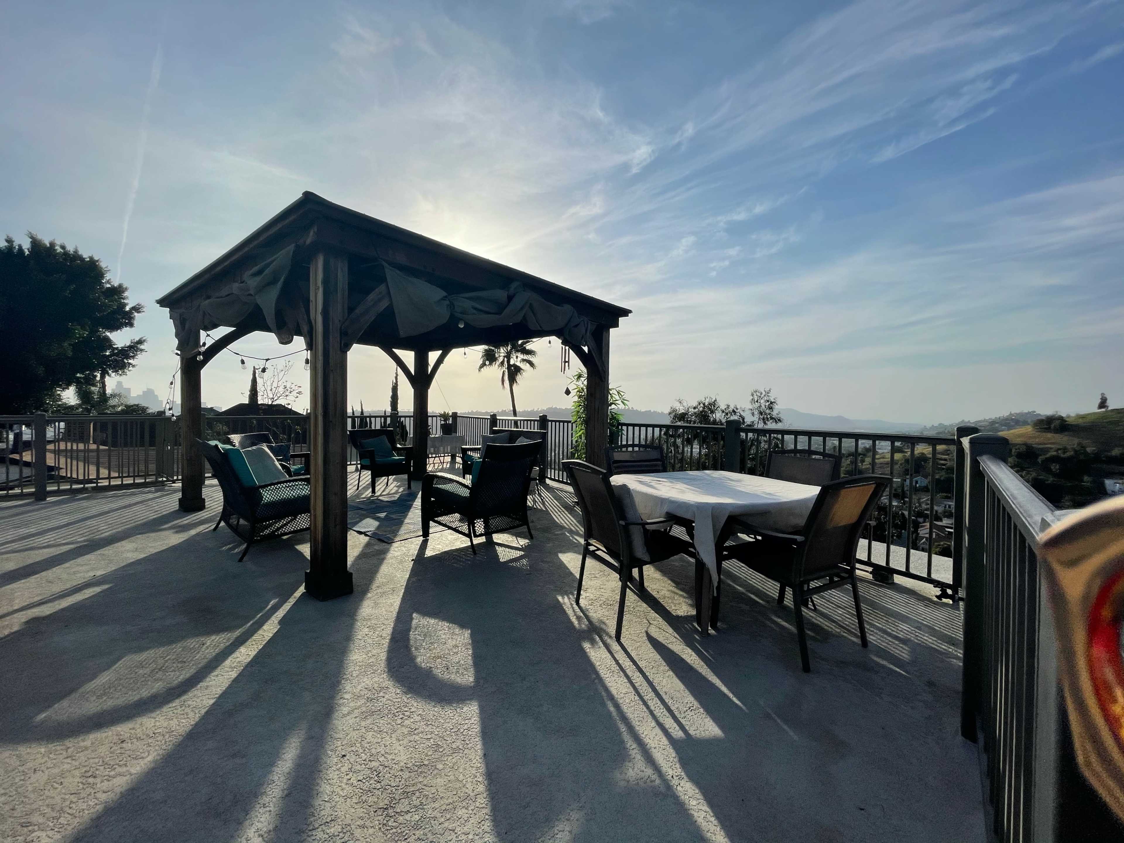 A wooden pergola with seating and tables is positioned on a terrace, casting long shadows against a backdrop of a clear sky.