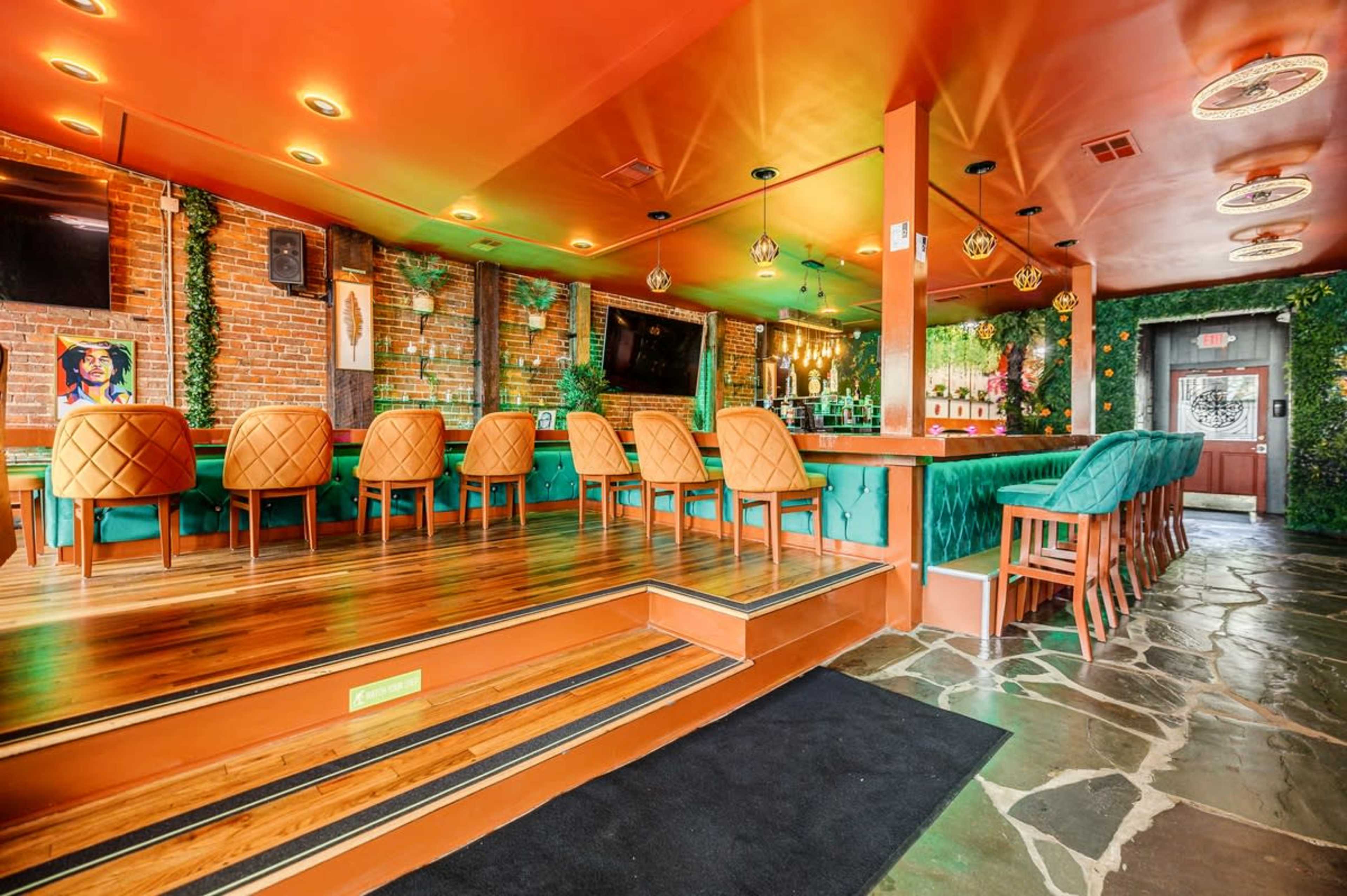 A vibrant bar interior with orange walls, green seating, and wooden flooring, featuring a mix of high and low seating arrangements.