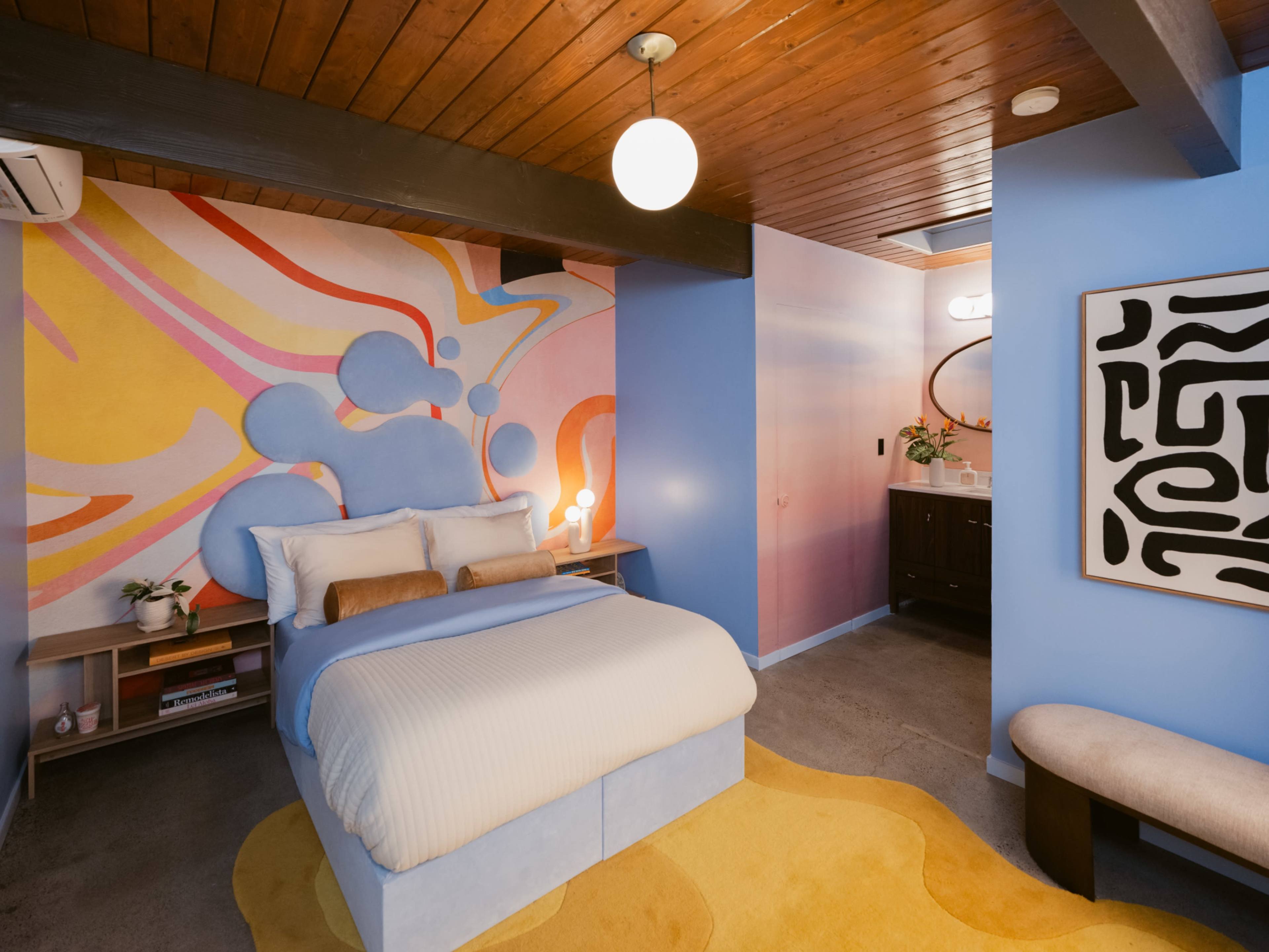 The image shows a modern bedroom featuring a bed with white linens, a colorful abstract mural on the wall, and a mix of wooden and painted surfaces.