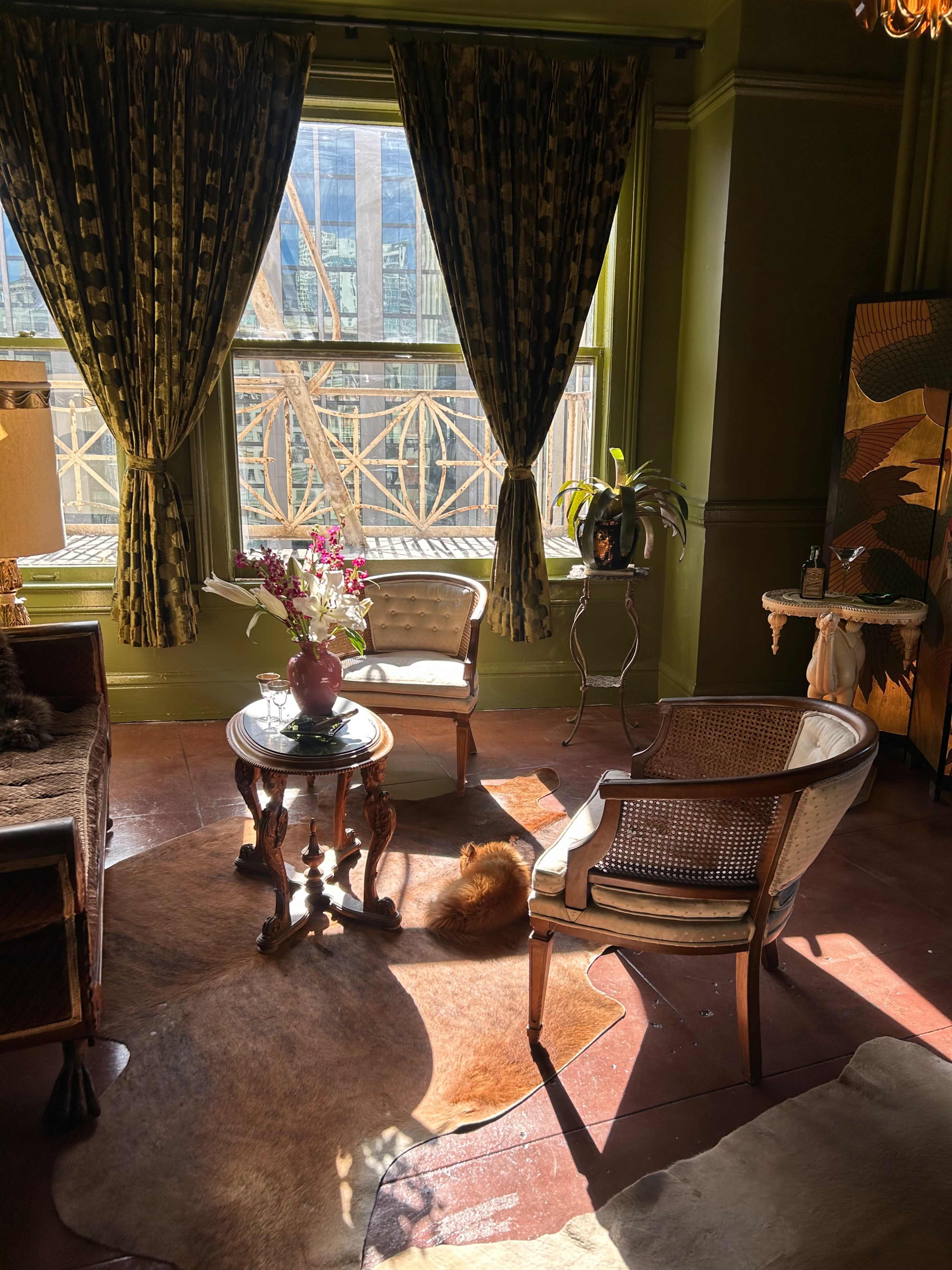 The room features two wicker chairs and a small wooden table on a cowhide rug, illuminated by sunlight streaming through large windows adorned with curtains.