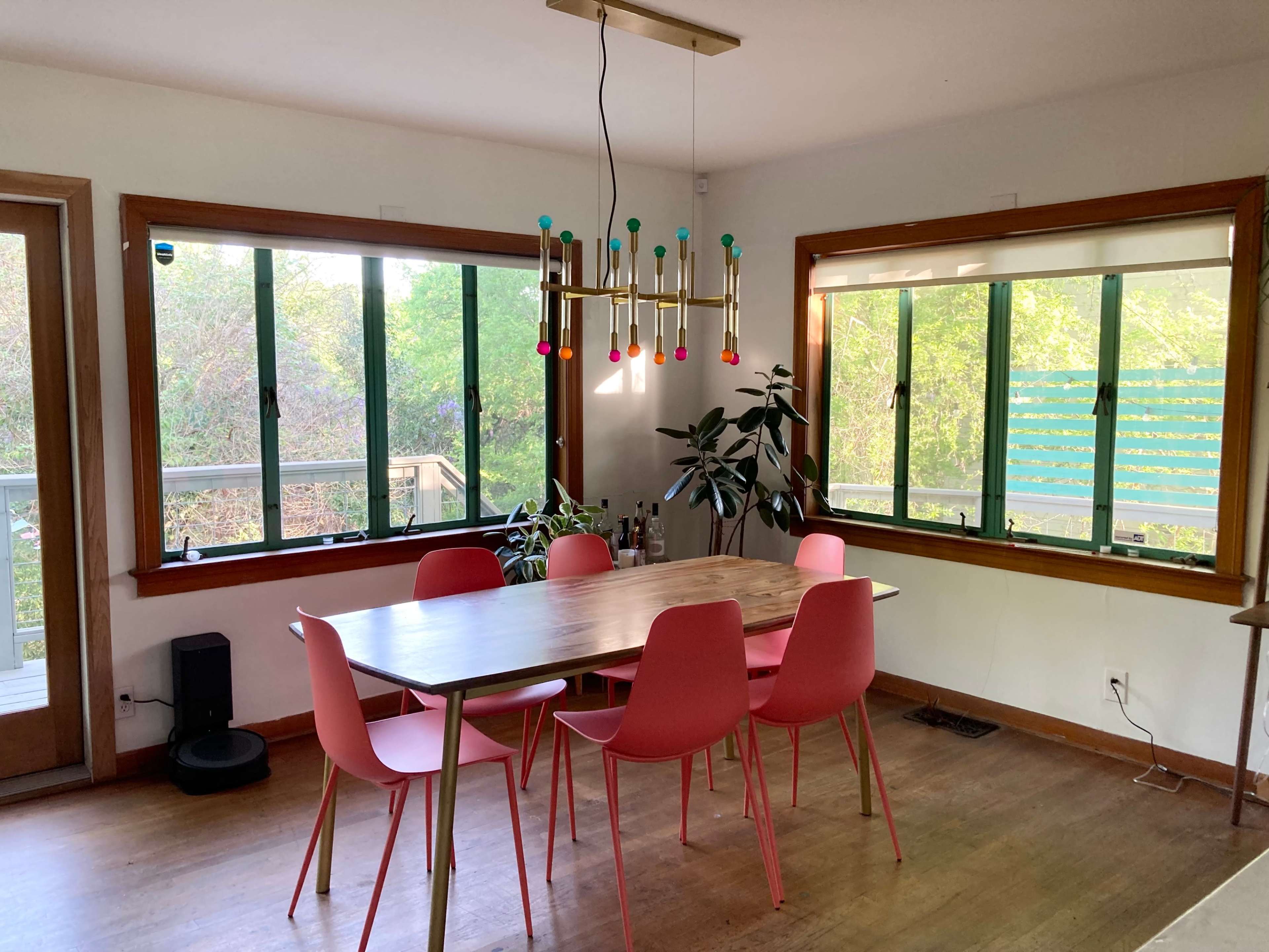A dining area features a wooden table surrounded by pink chairs, with large windows offering a view of greenery outside.