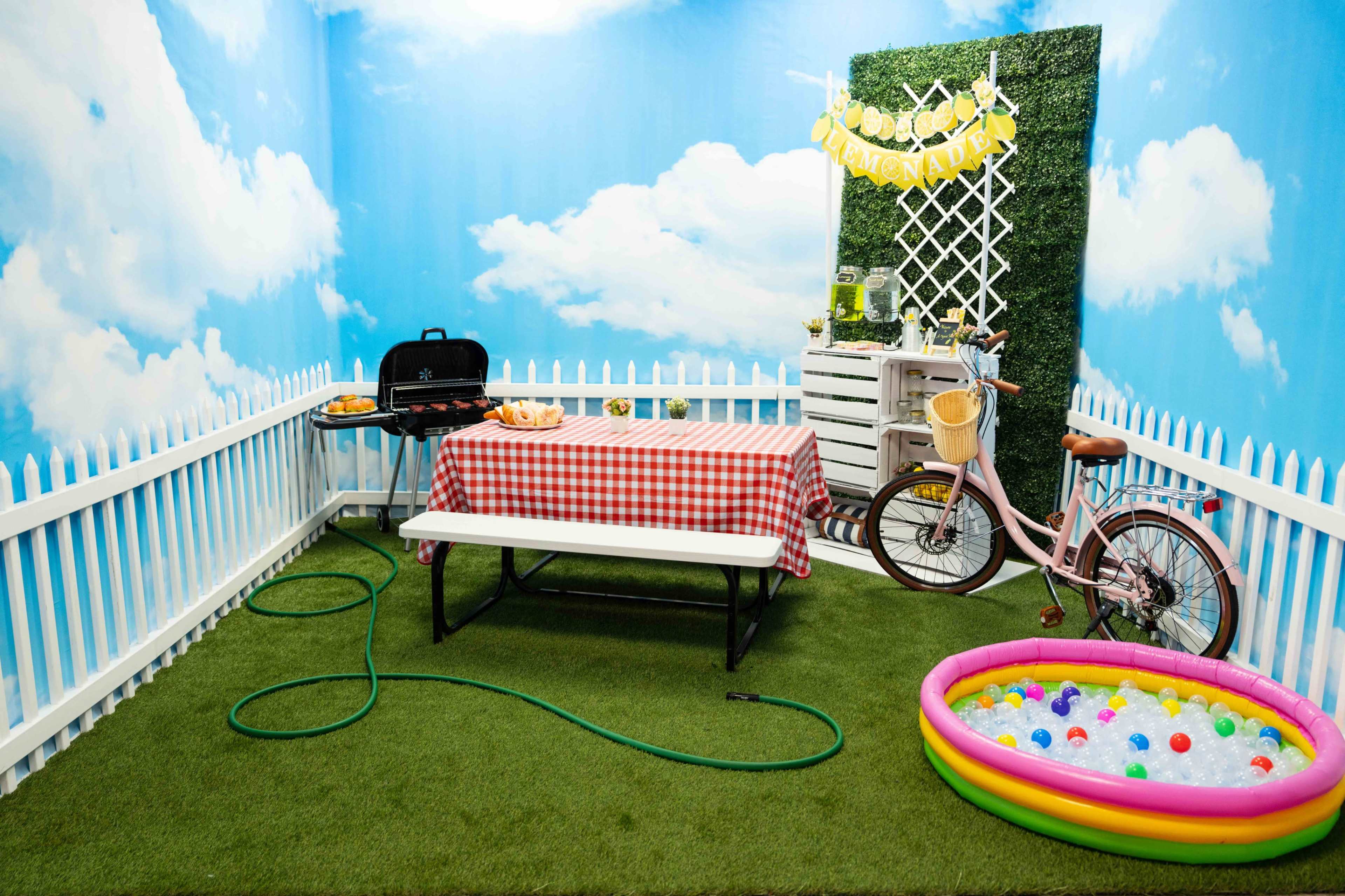 The scene features a small outdoor area with a picnic table covered in a red-and-white checked tablecloth, a grill, a bicycle, and a colorful inflatable pool filled with balls.