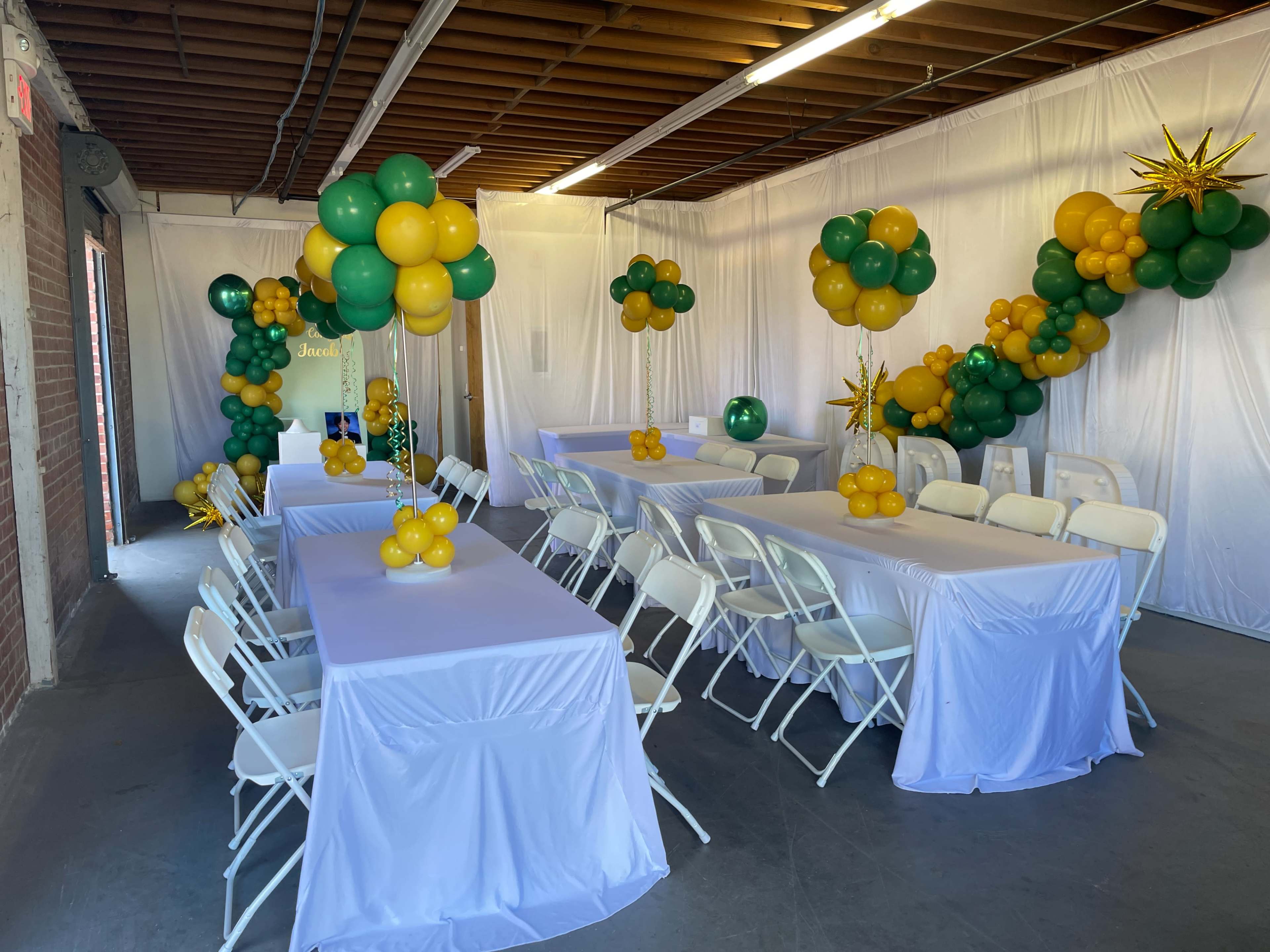 A decorated indoor space with white tables covered in tablecloths, surrounded by rows of folding chairs and adorned with yellow and green balloons in various arrangements.
