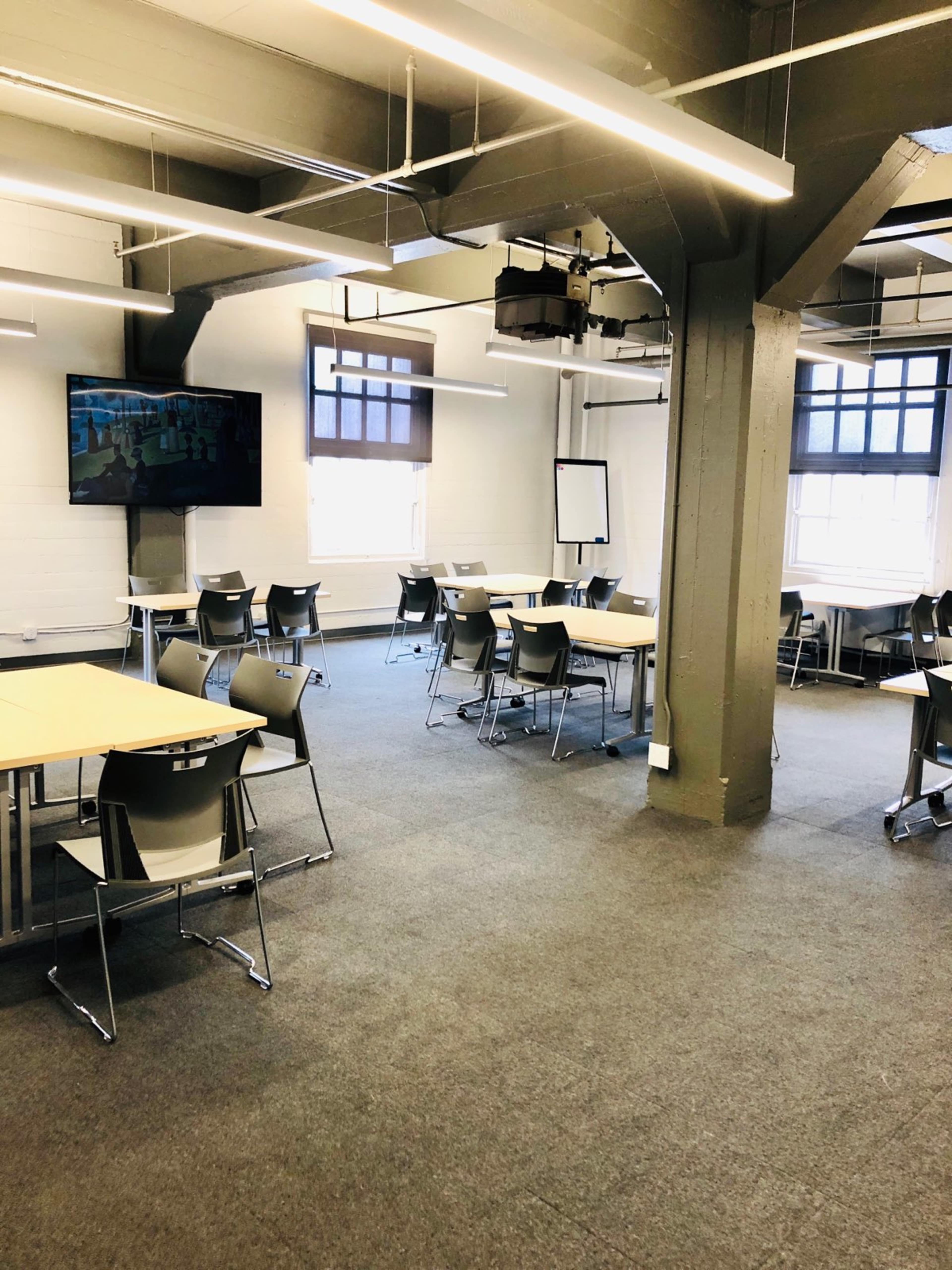 A modern classroom with several tables and chairs arranged neatly, and a large screen mounted on the wall.