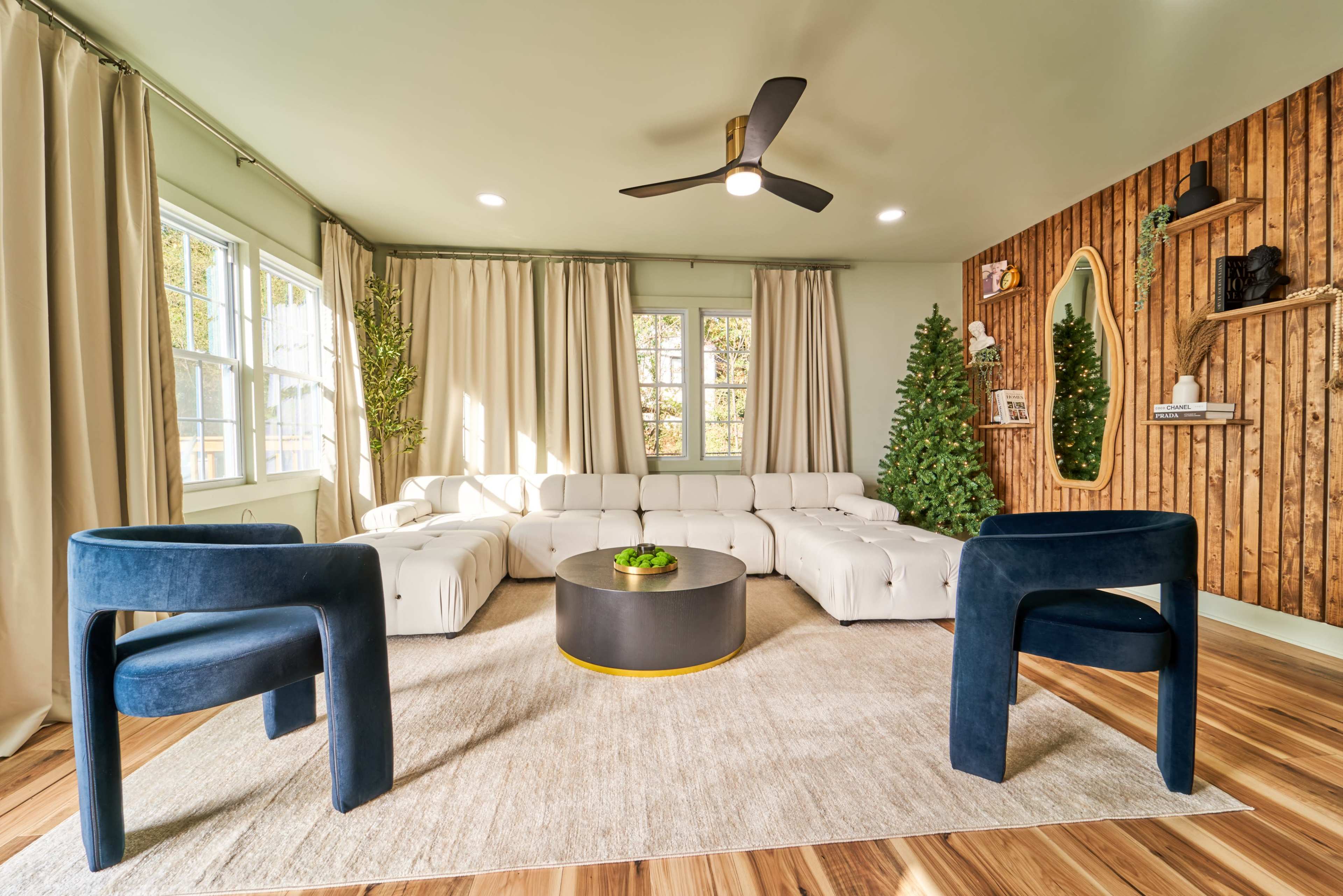 A modern living room features a white sectional sofa, two dark blue chairs, a round black coffee table, and floor-to-ceiling curtains, all set against a wooden accent wall adorned with decorative items.