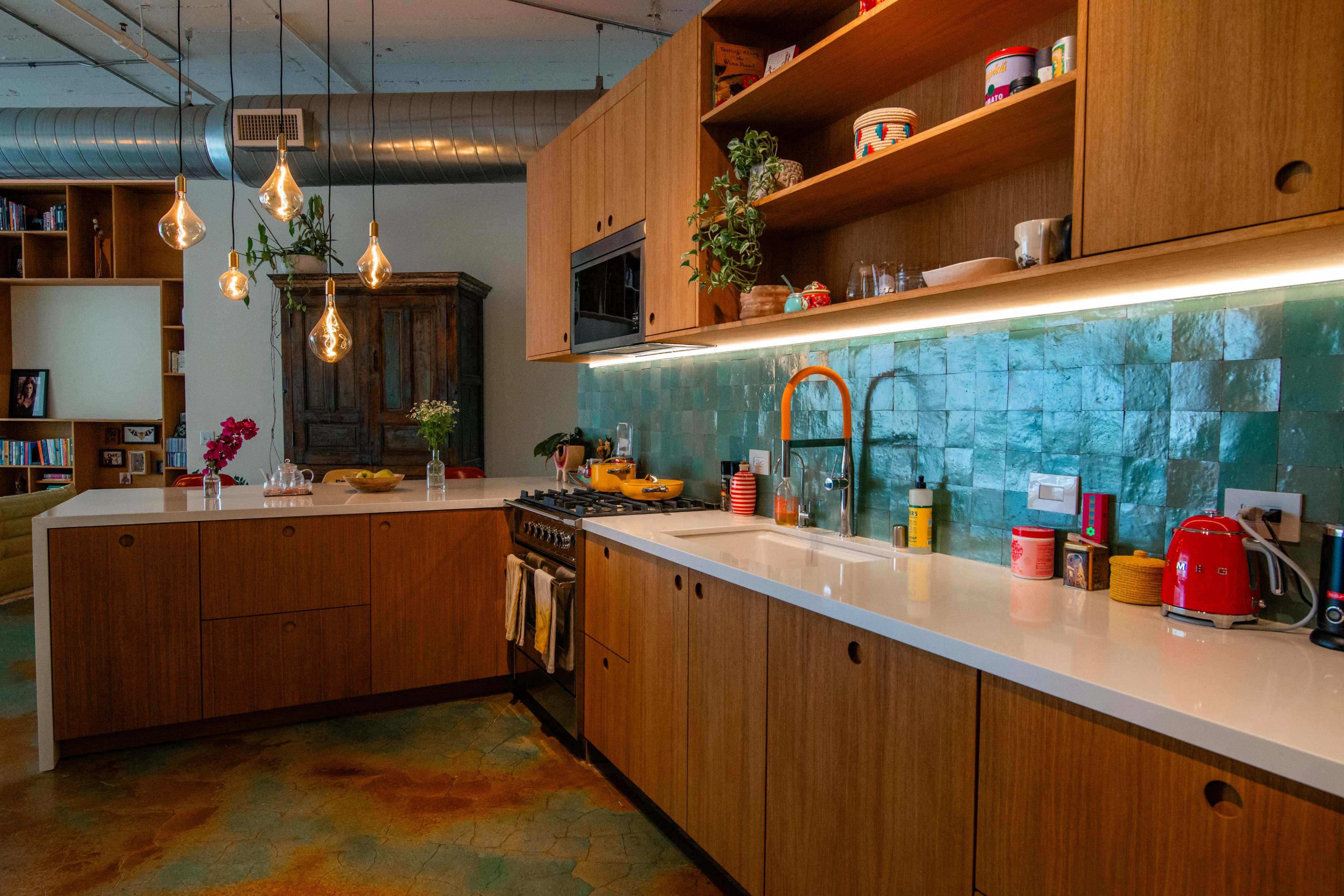 The image shows a modern kitchen featuring wooden cabinetry, a white countertop, colorful decor, and pendant lights hanging above the workspace.