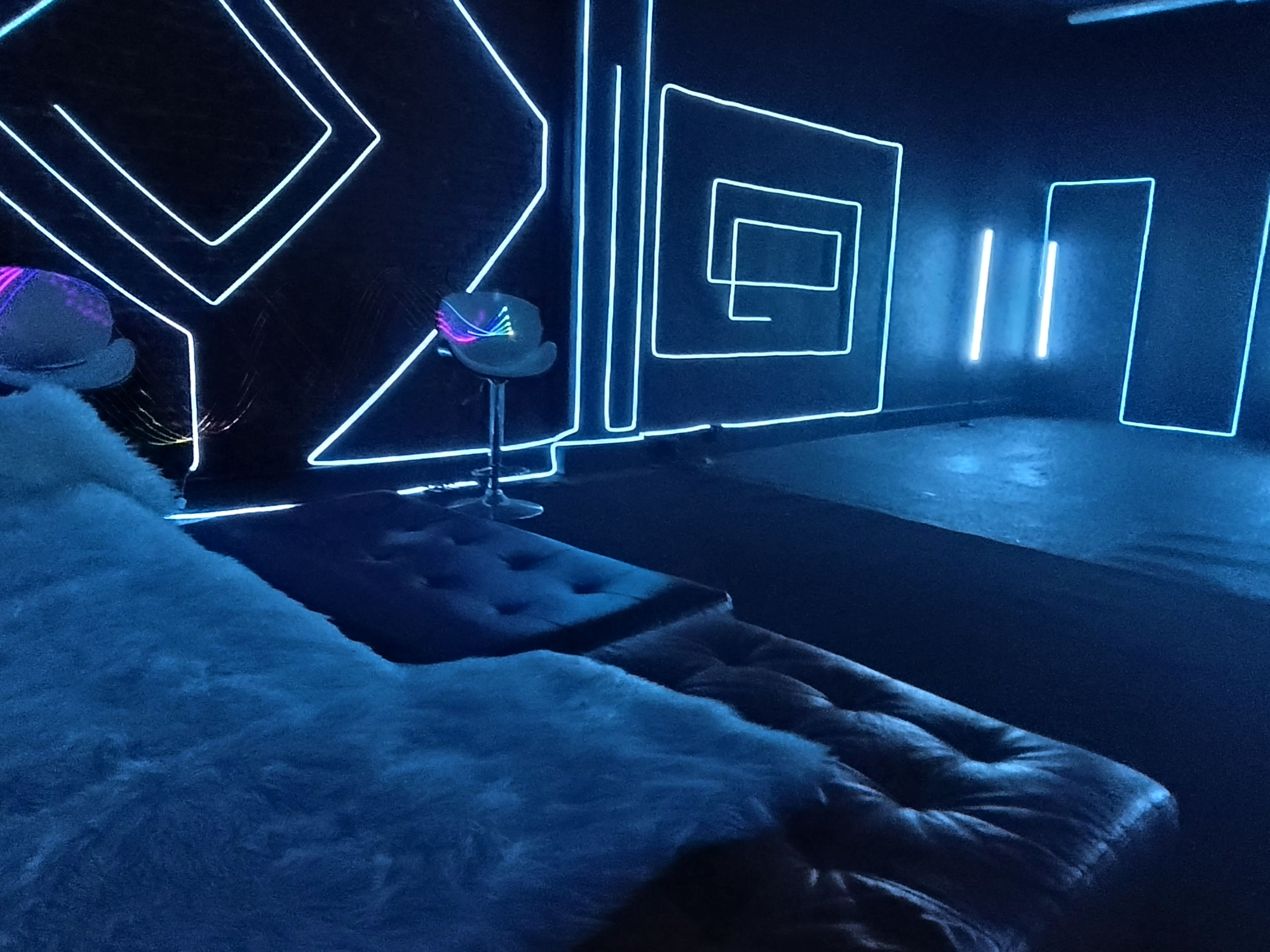 The image shows a dimly lit room with neon blue geometric lines on the walls, a plush sofa, and modern chairs illuminated by colorful lights.