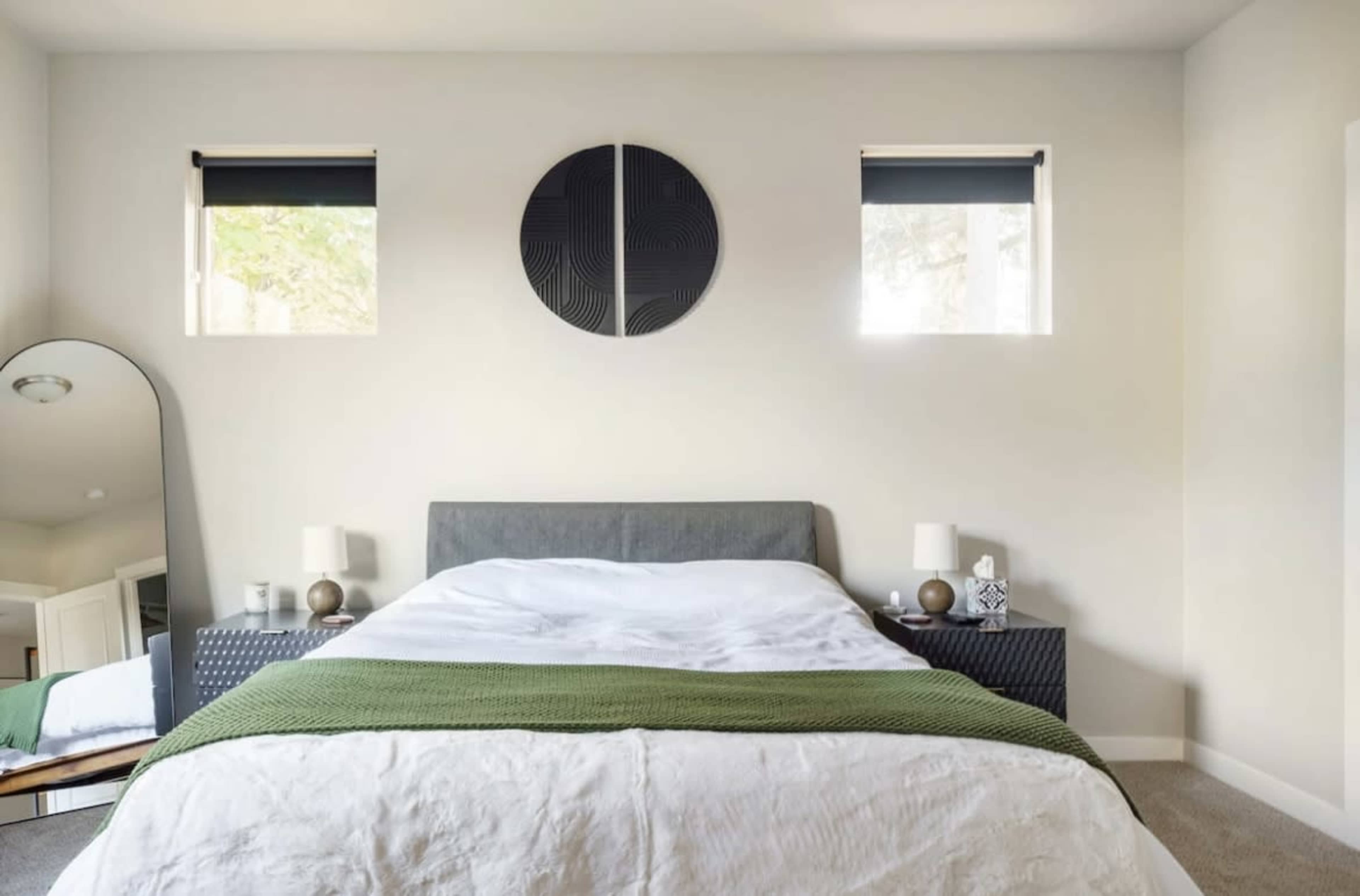A neatly arranged bedroom with a bed covered in white bedding, a green throw blanket, and two windows with shades above nightstands on either side.