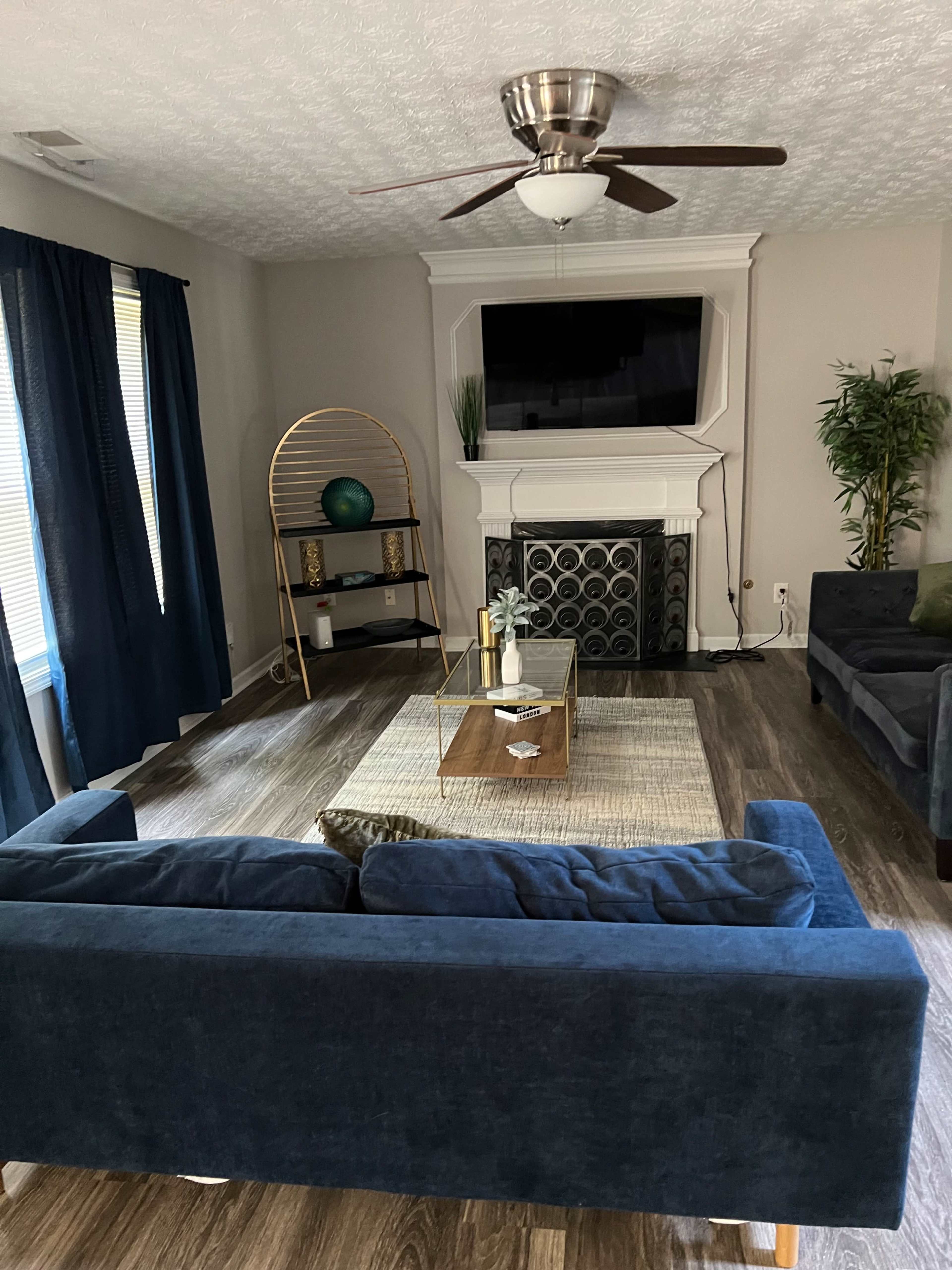 The living room features a blue sofa, a coffee table on a textured rug, and a media console beneath a mounted TV, with large windows dressed in navy curtains.