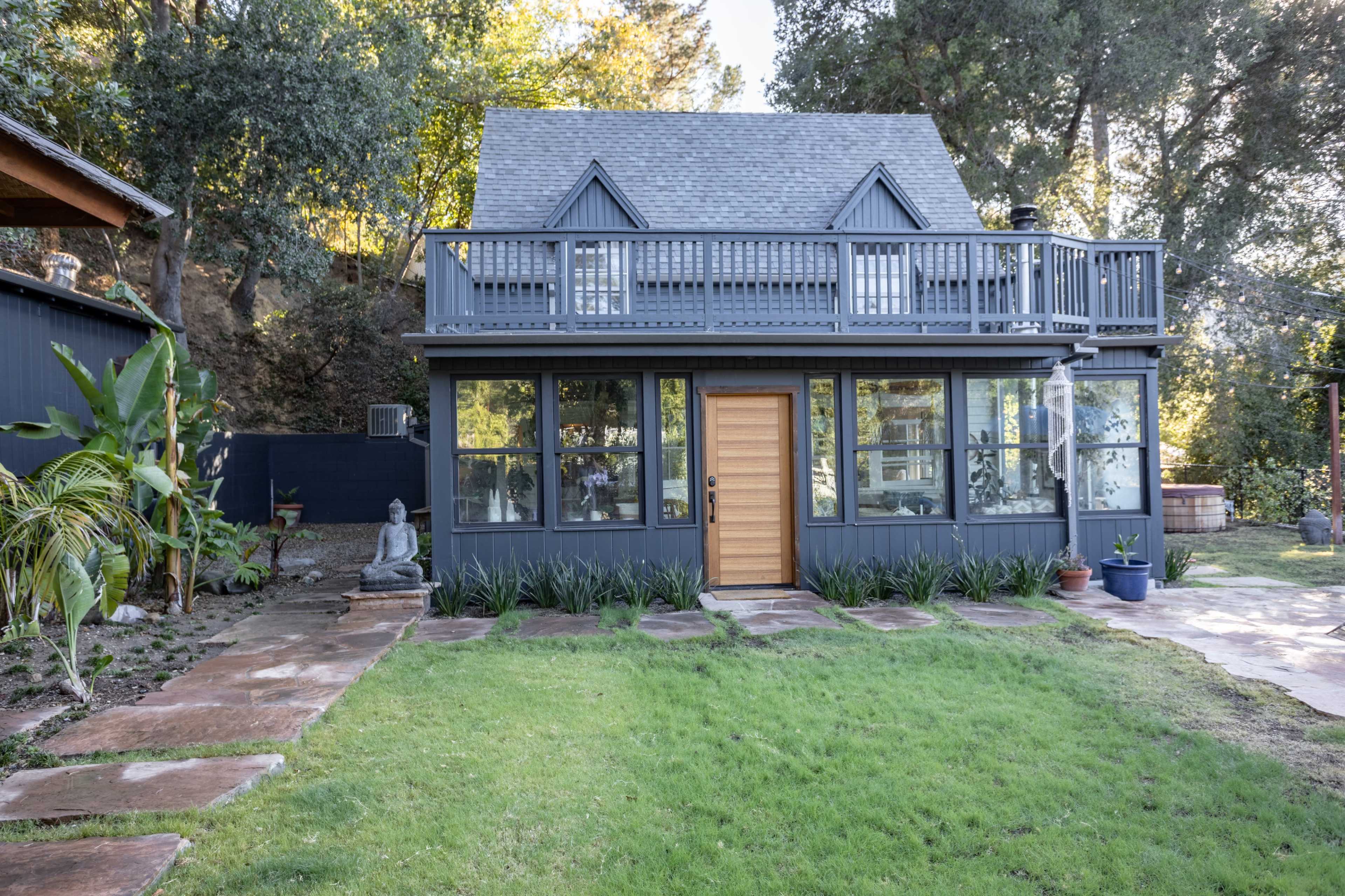 A two-story gray house with a wooden front door, large windows, and a balcony surrounded by a lawn and landscaped garden.