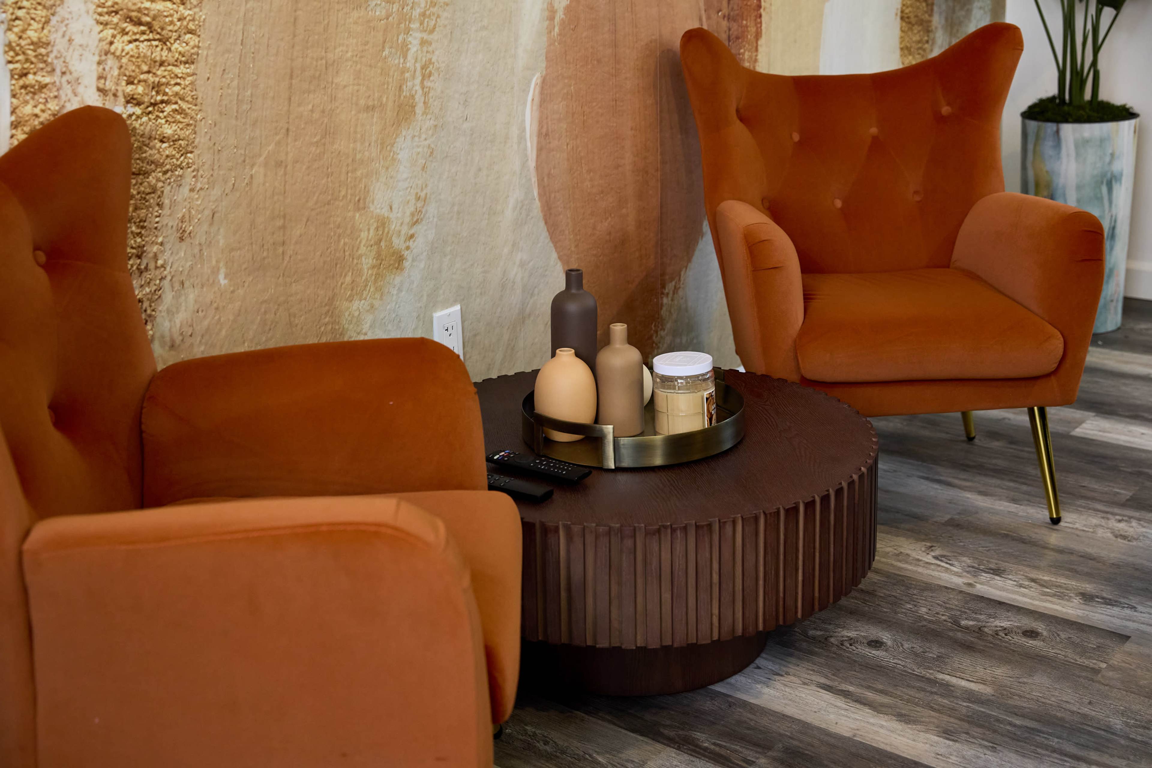 The image features two orange armchairs flanking a round wooden coffee table with decorative vases and a tray, set against a textured wall.