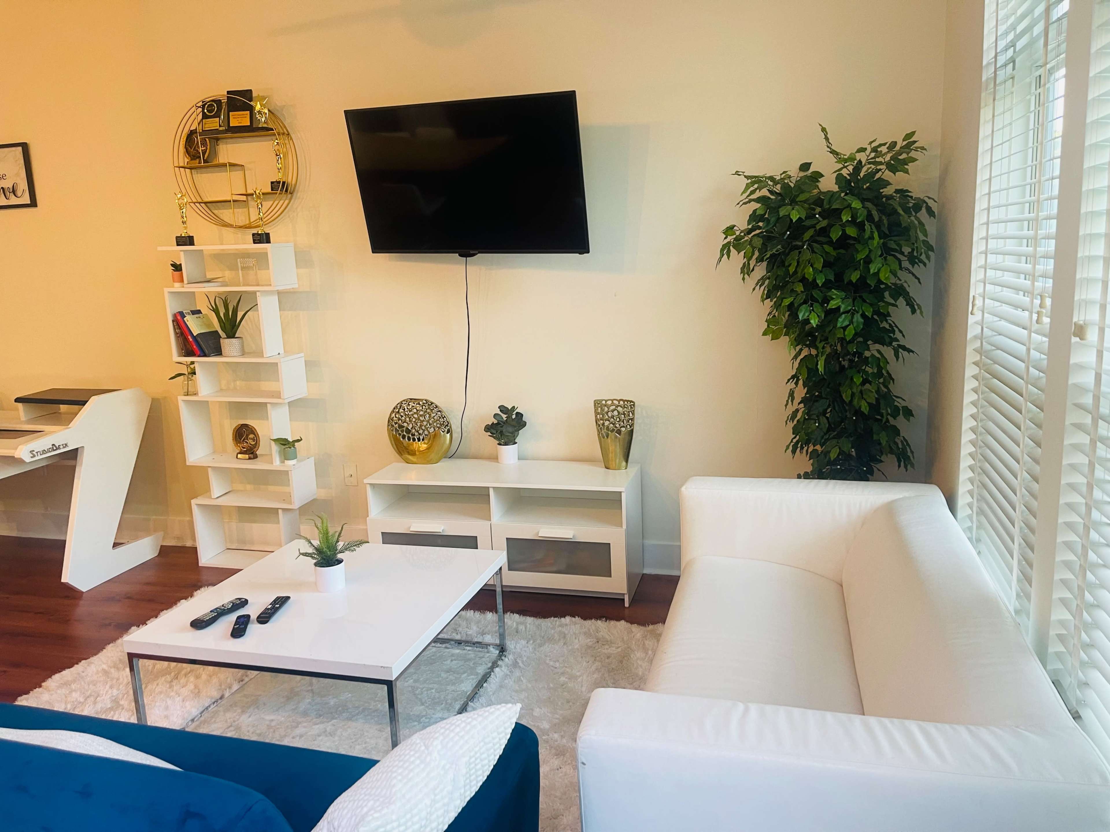 A living room with a white sofa, a coffee table, a TV mounted on the wall, and a shelf with decorative items and plants.