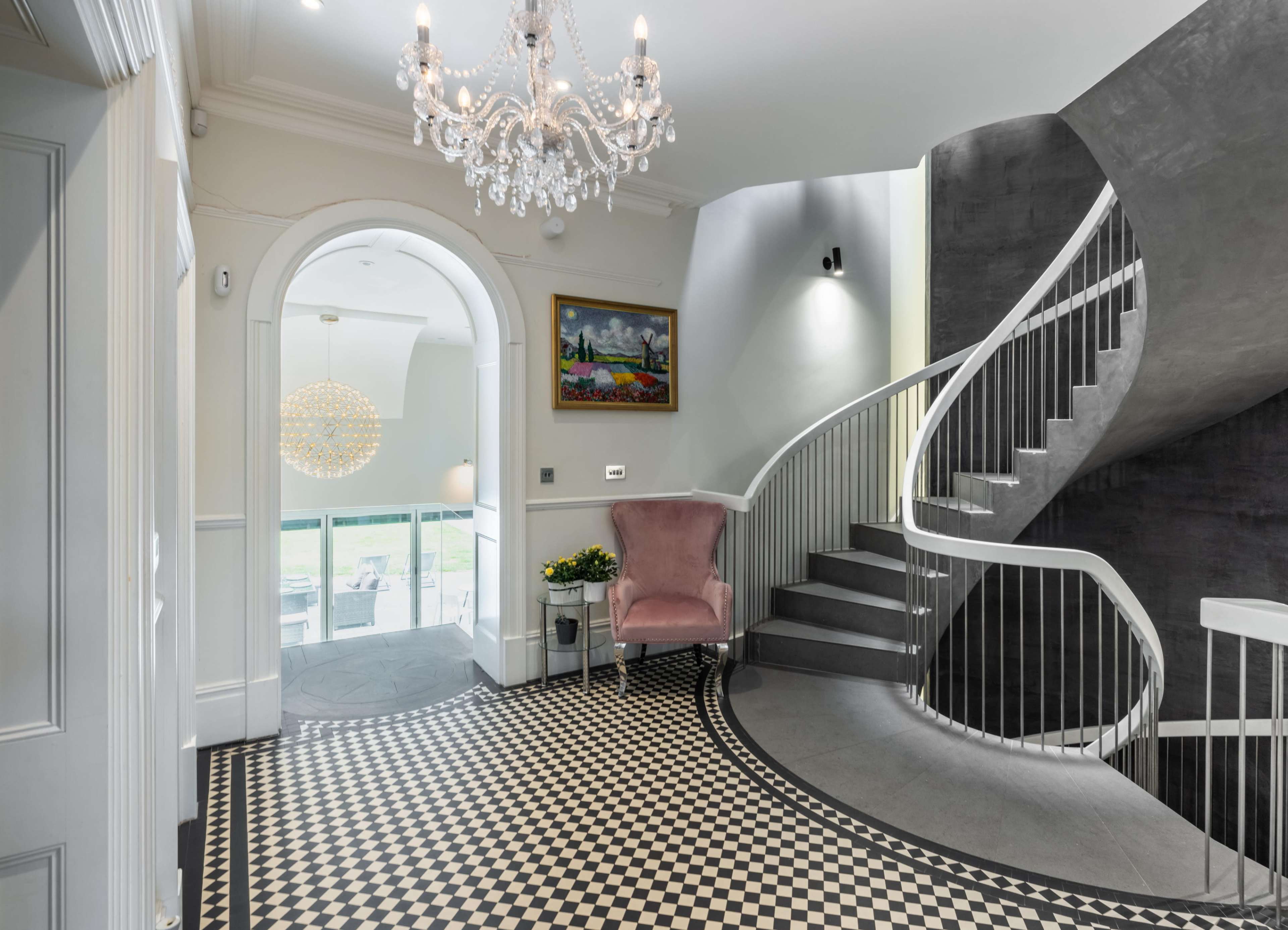 A curved staircase is featured in an entryway with a checkered floor and a chandelier, complemented by a decorative chair and artwork on the wall.