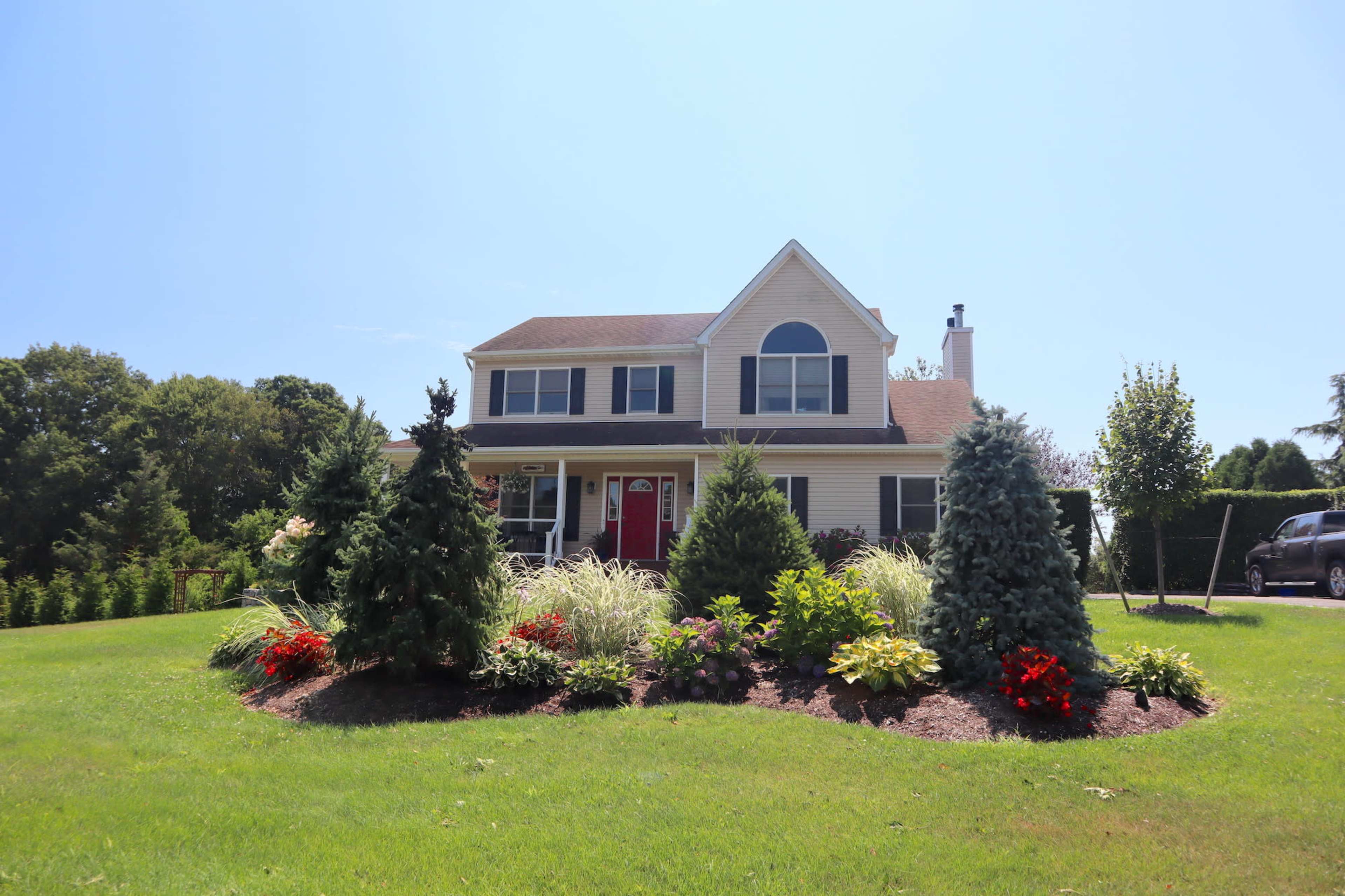 A two-story house with a red door is surrounded by well-maintained landscaping featuring flowering plants and evergreen trees.