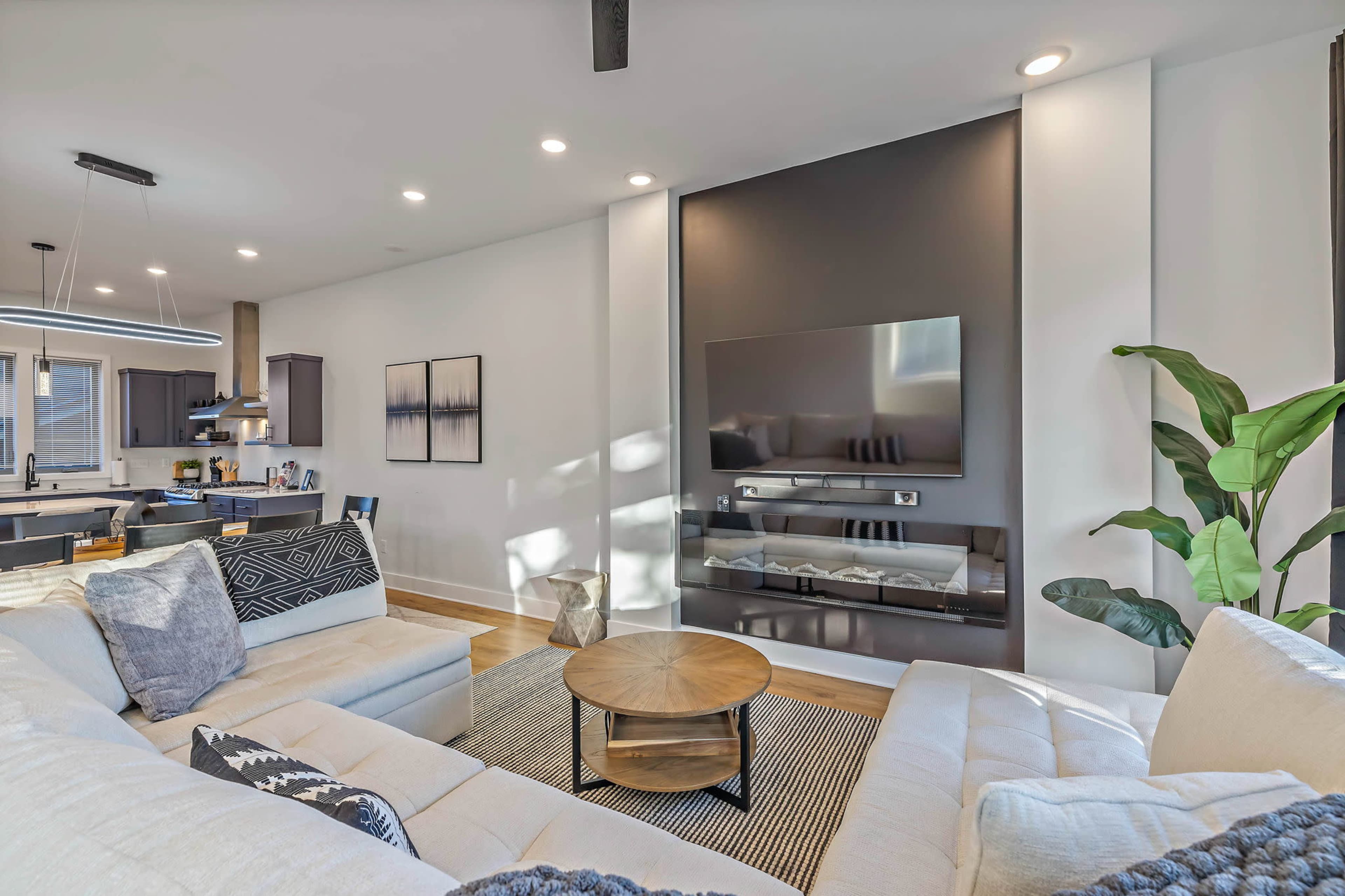 The image shows a modern living room featuring a sectional sofa, a round coffee table, and a wall-mounted television, with a kitchen visible in the background.