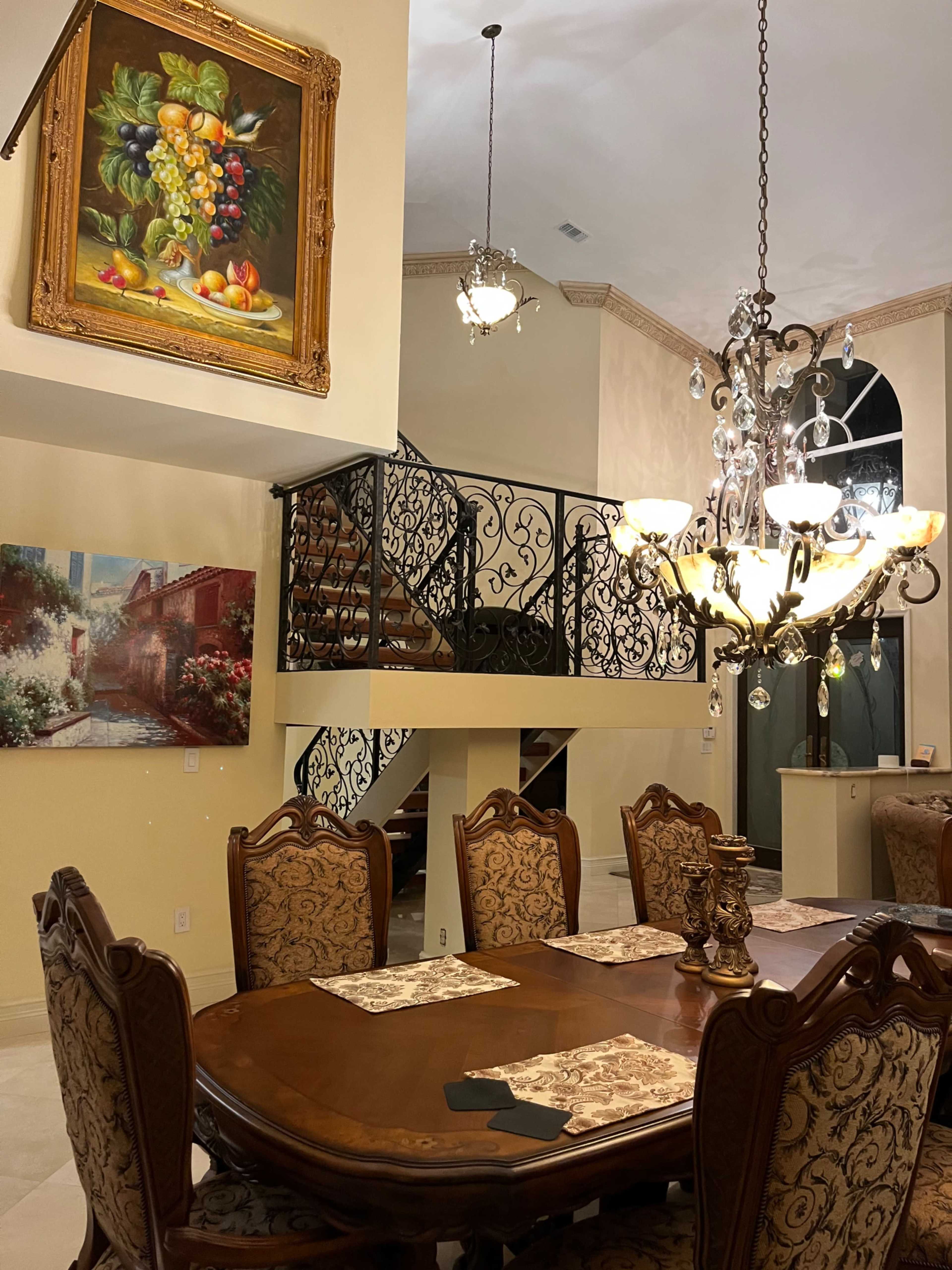 A spacious dining area with a wooden table set for dinner, a decorative chandelier above, and a staircase adorned with an ornate iron railing in the background.