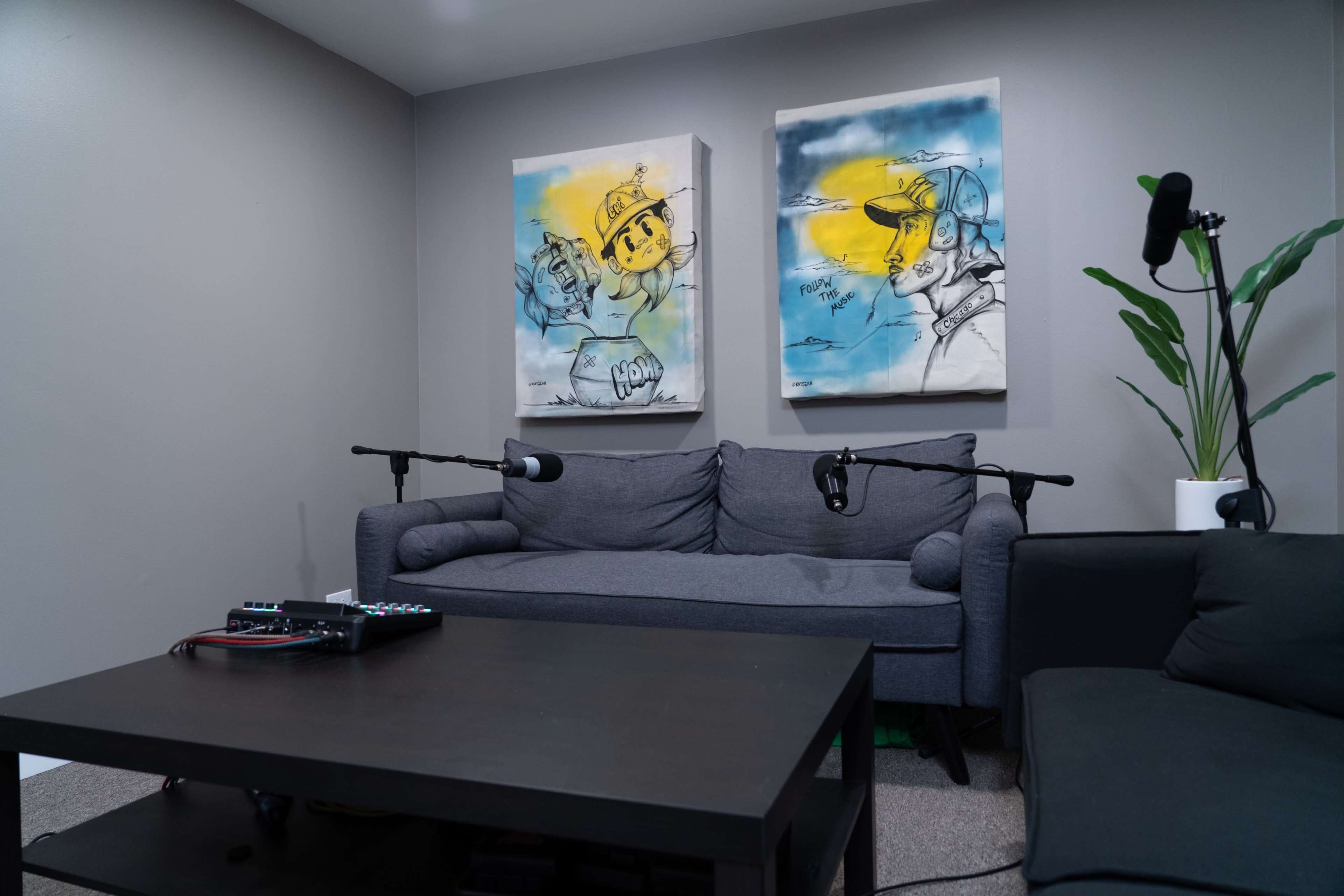 A modern living space with two framed artworks on the wall, a gray couch, a black coffee table with equipment, and plants in the corners.