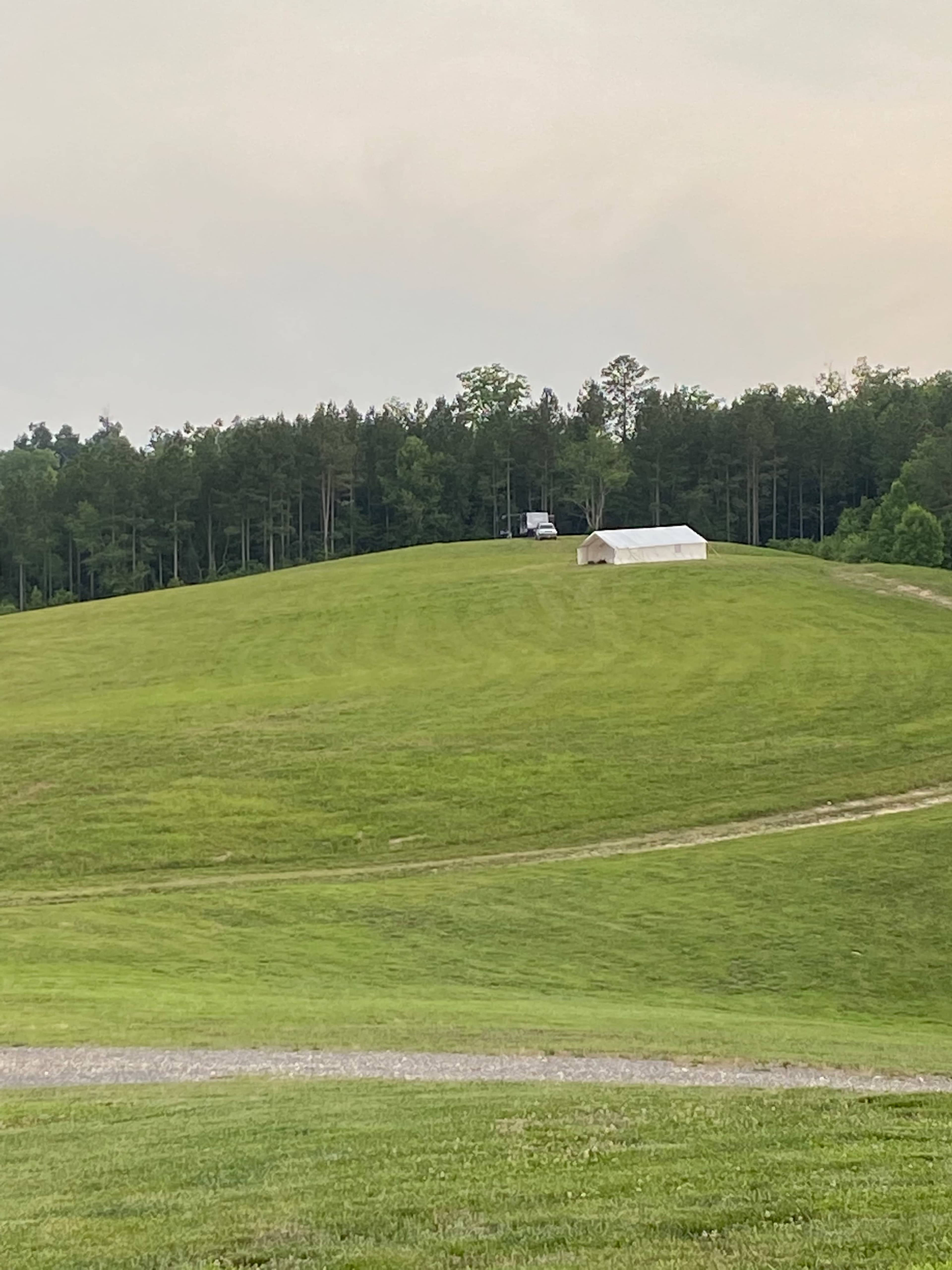 A white tent is set up on a grassy hill surrounded by a tree line.