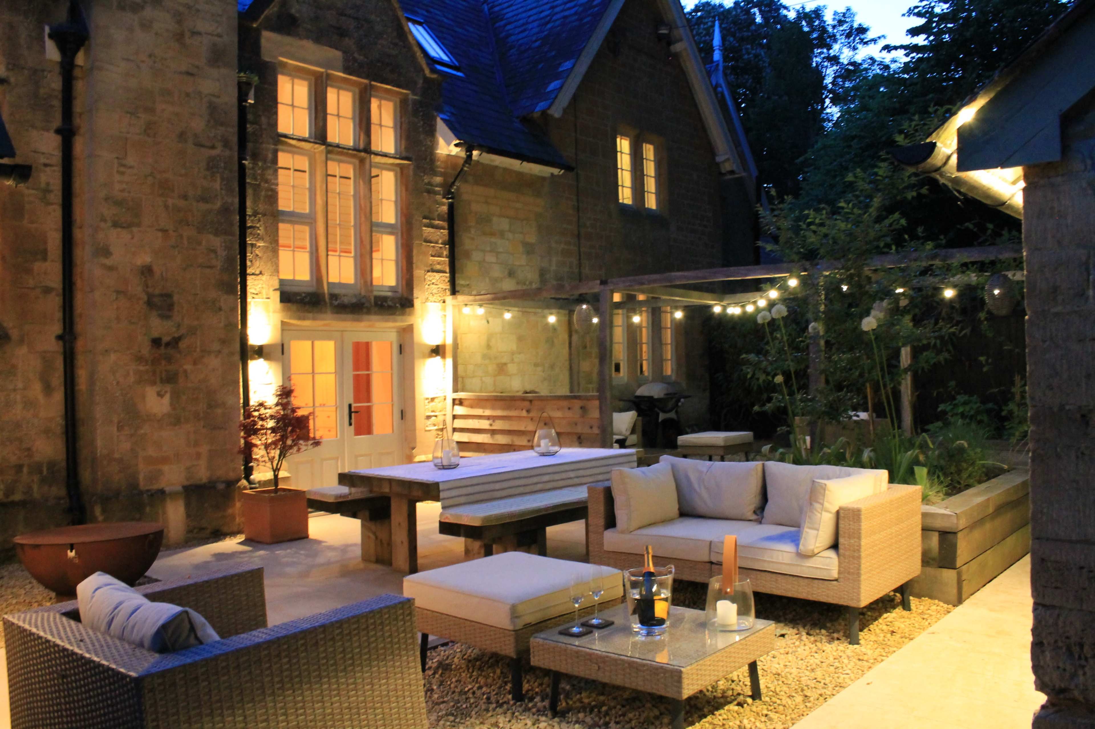 A beautifully lit outdoor patio features a seating area with wicker furniture, a large wooden table, and string lights surrounding a garden.