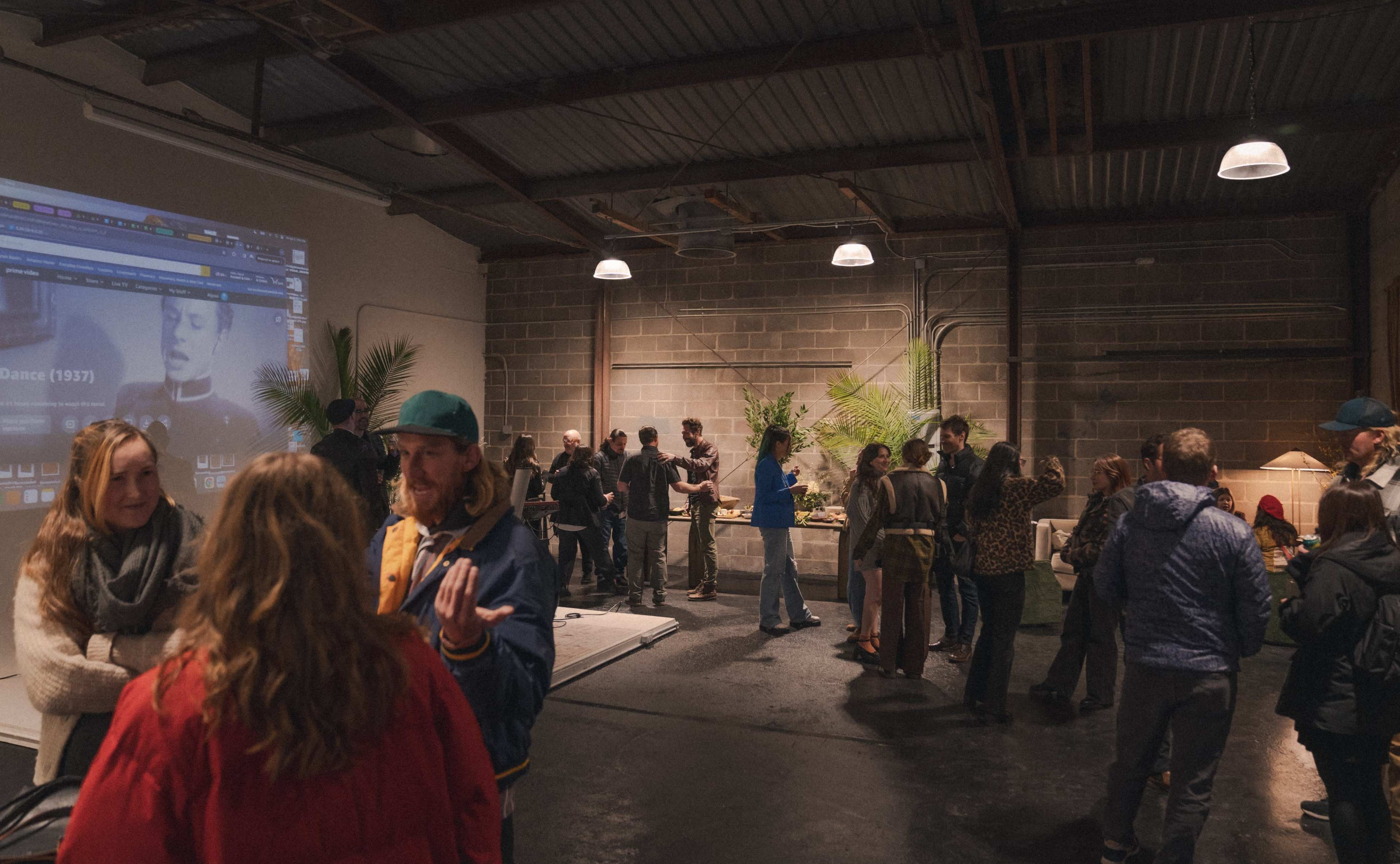 A group of people gathers in an industrial-style space with a large screen displaying video content, while others engage in conversation amidst plants and minimal furnishings.