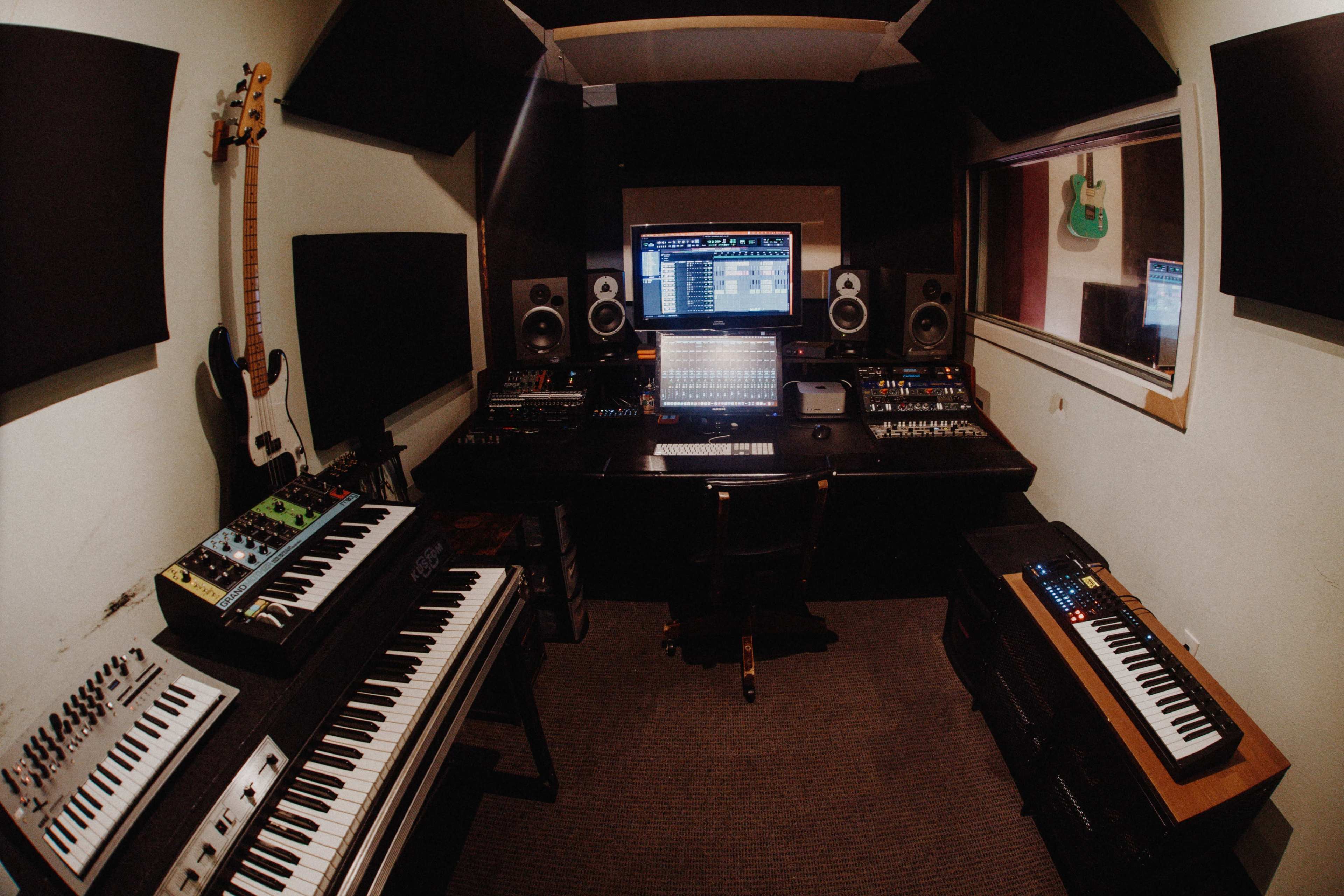 A music production studio features a central mixing console, synthesizers, and various audio equipment, with soundproof panels on the walls.
