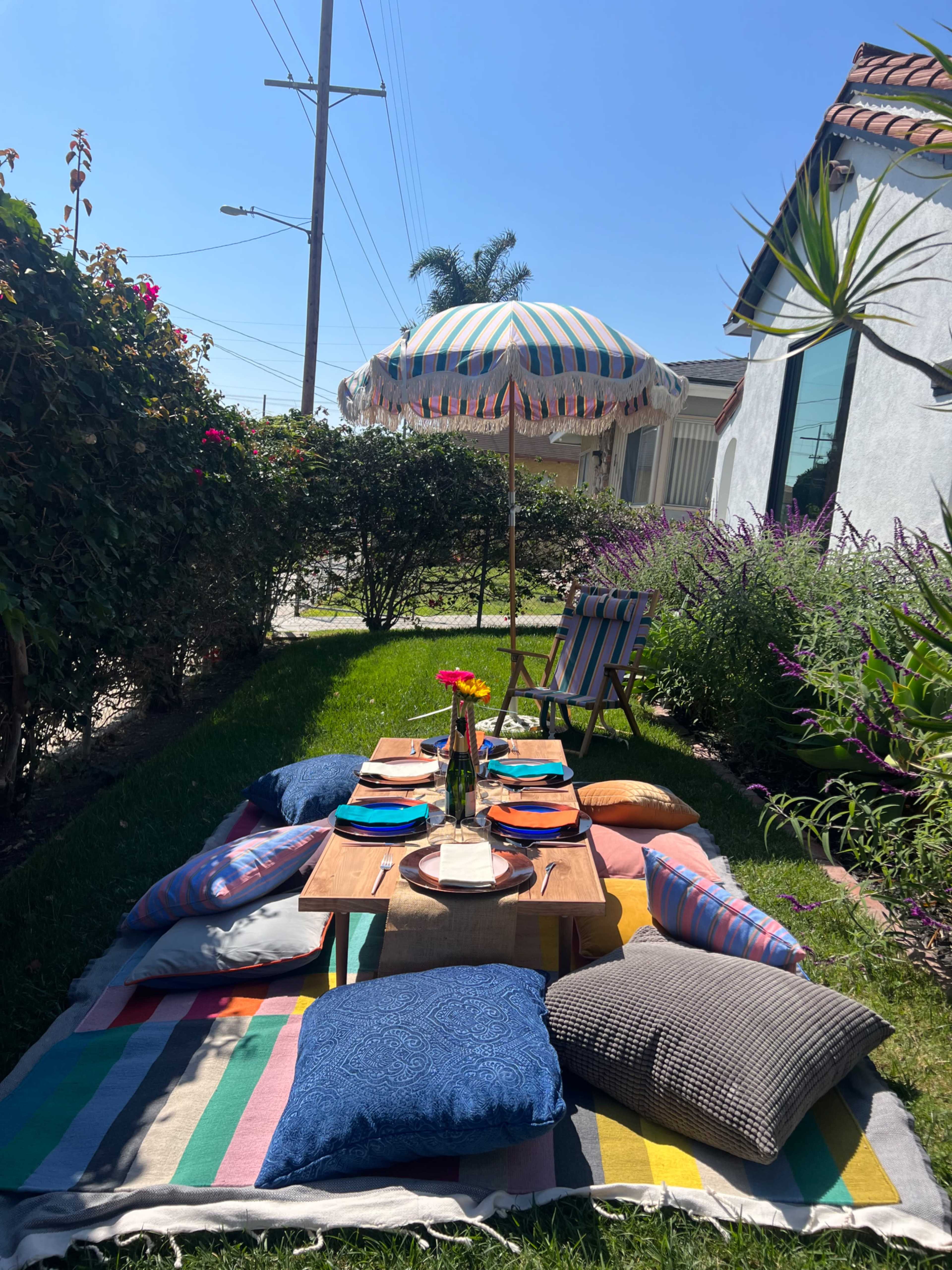 A picnic setting is arranged on a striped blanket under a decorative umbrella, surrounded by greenery and colorful cushions.