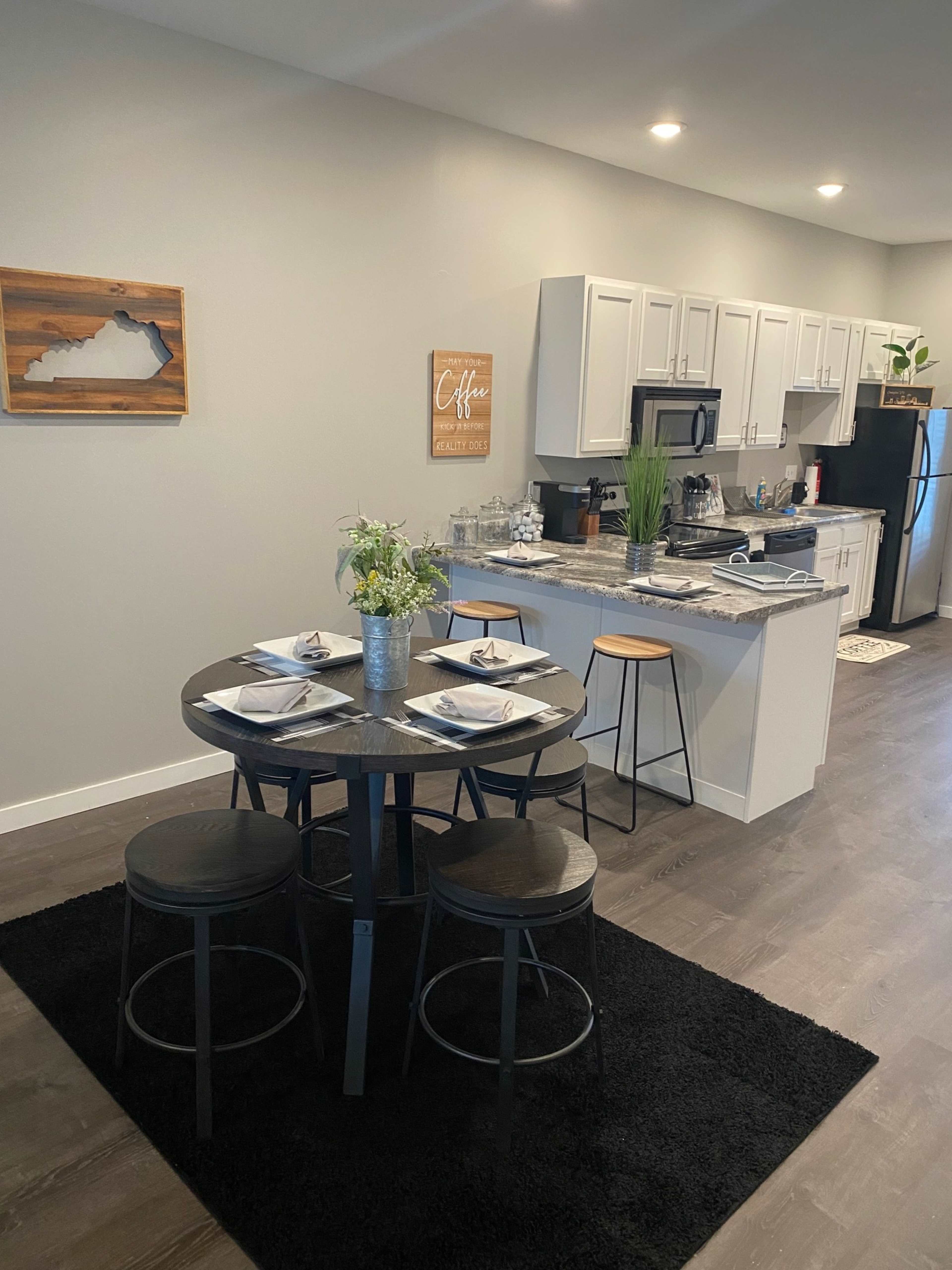 A modern dining area with a round table set for four, surrounded by black stools, adjacent to a kitchen with white cabinets and appliances.