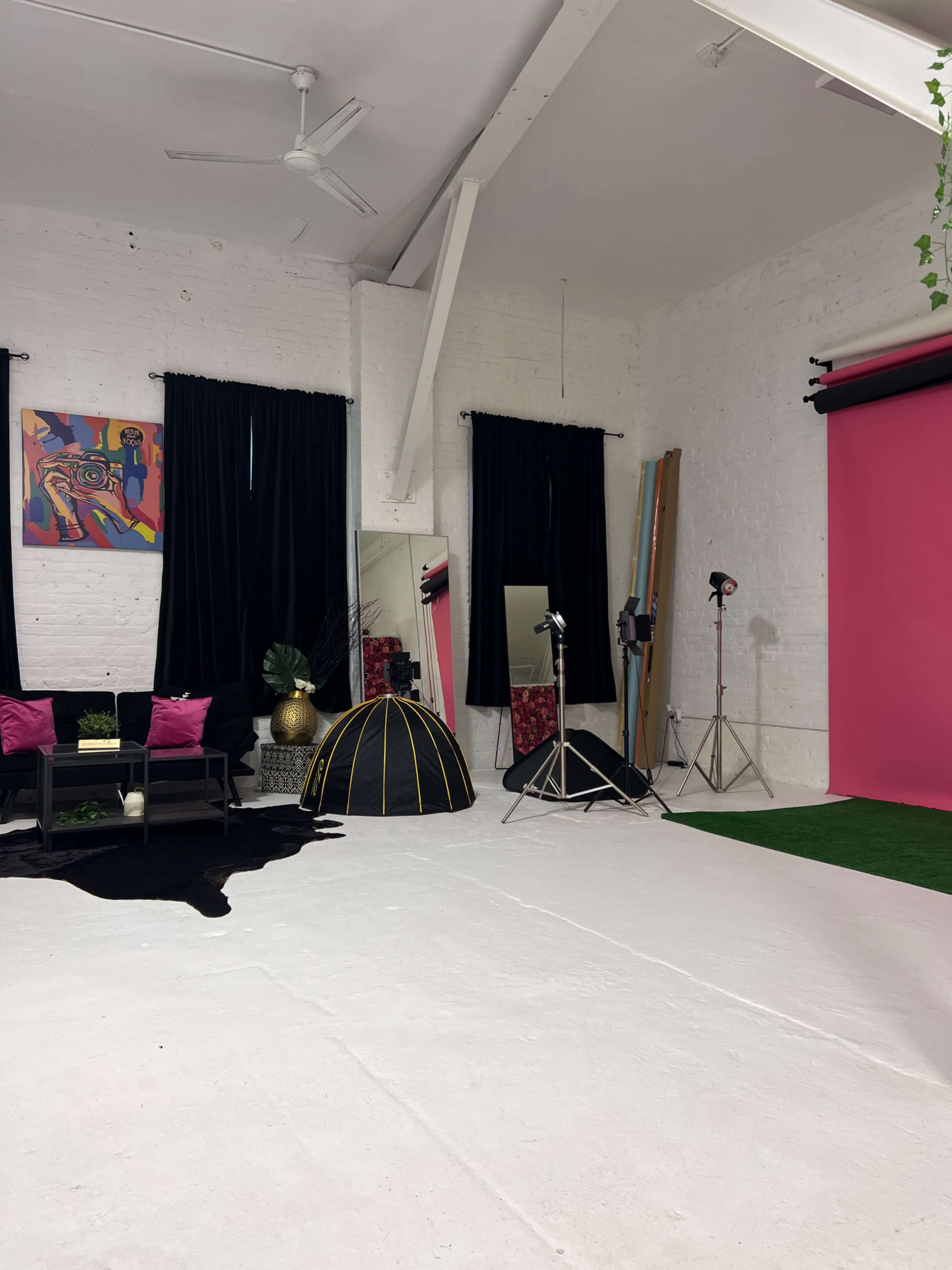 A photography studio with a pink backdrop, black curtains, a seating area, and various lighting equipment.