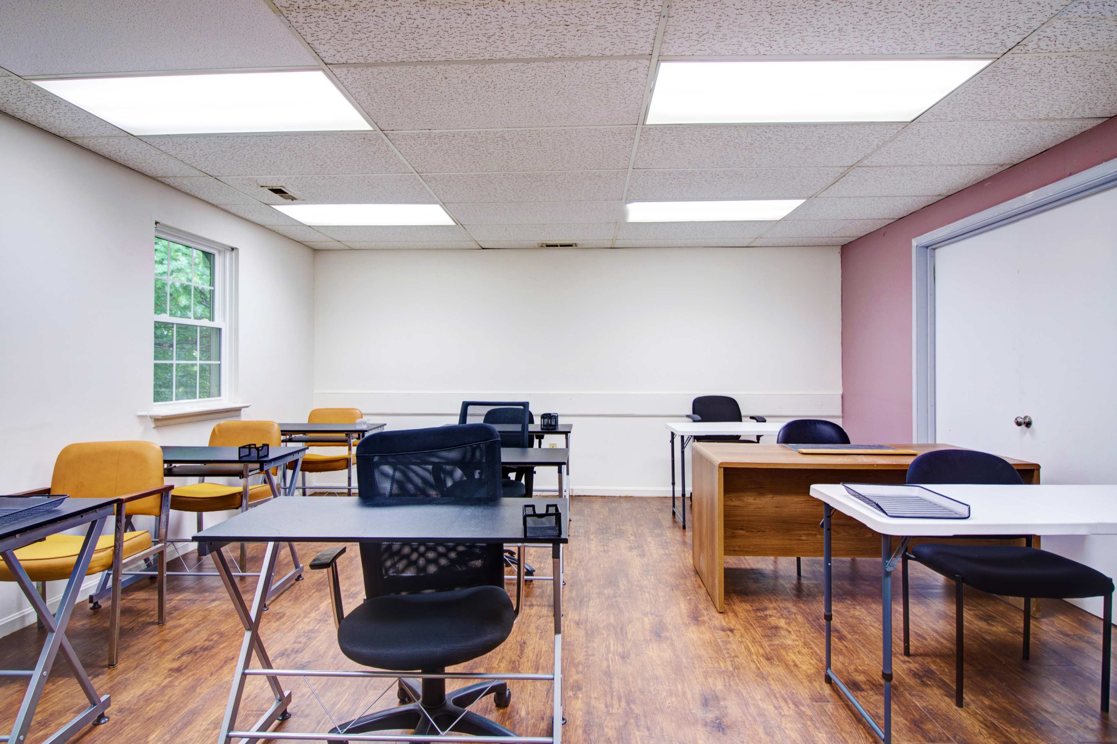 A classroom with several desks and chairs arranged in a mostly empty space, featuring a combination of dark and light furniture against a neutral-colored wall.