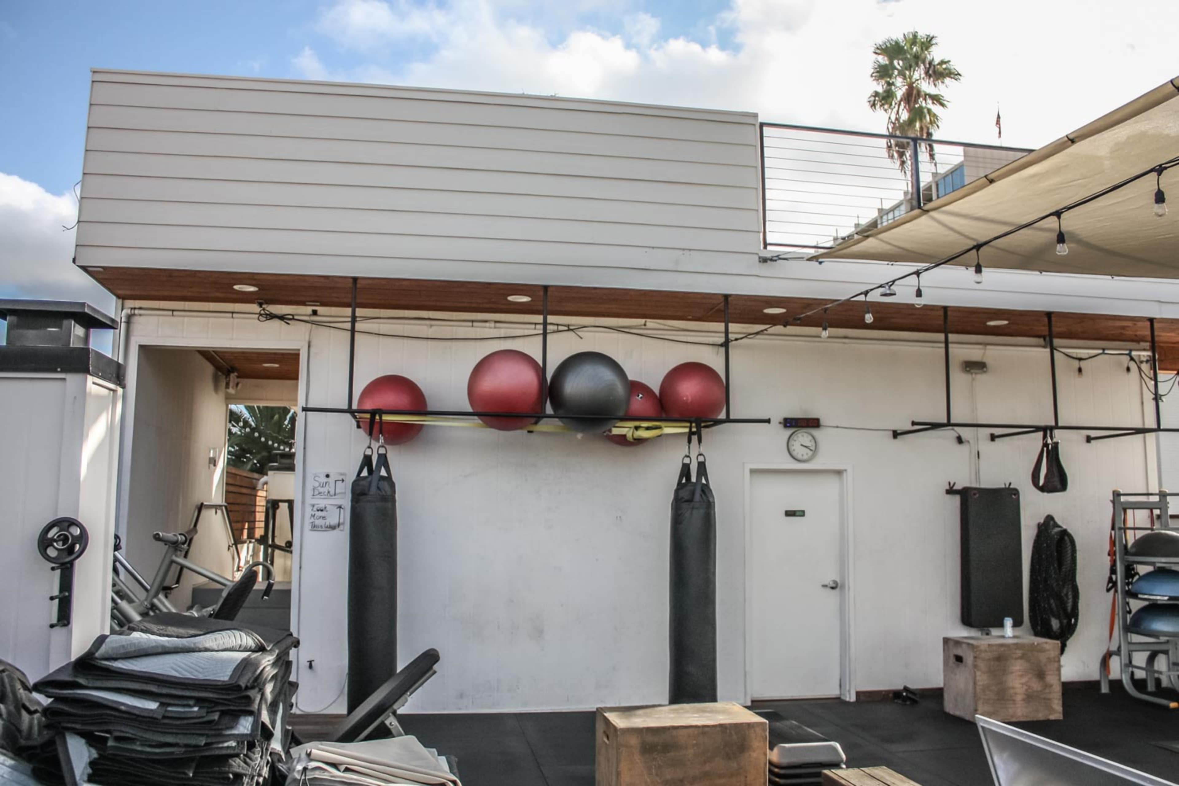 An outdoor gym area with exercise balls, punching bags, and various fitness equipment arranged against a white wall.