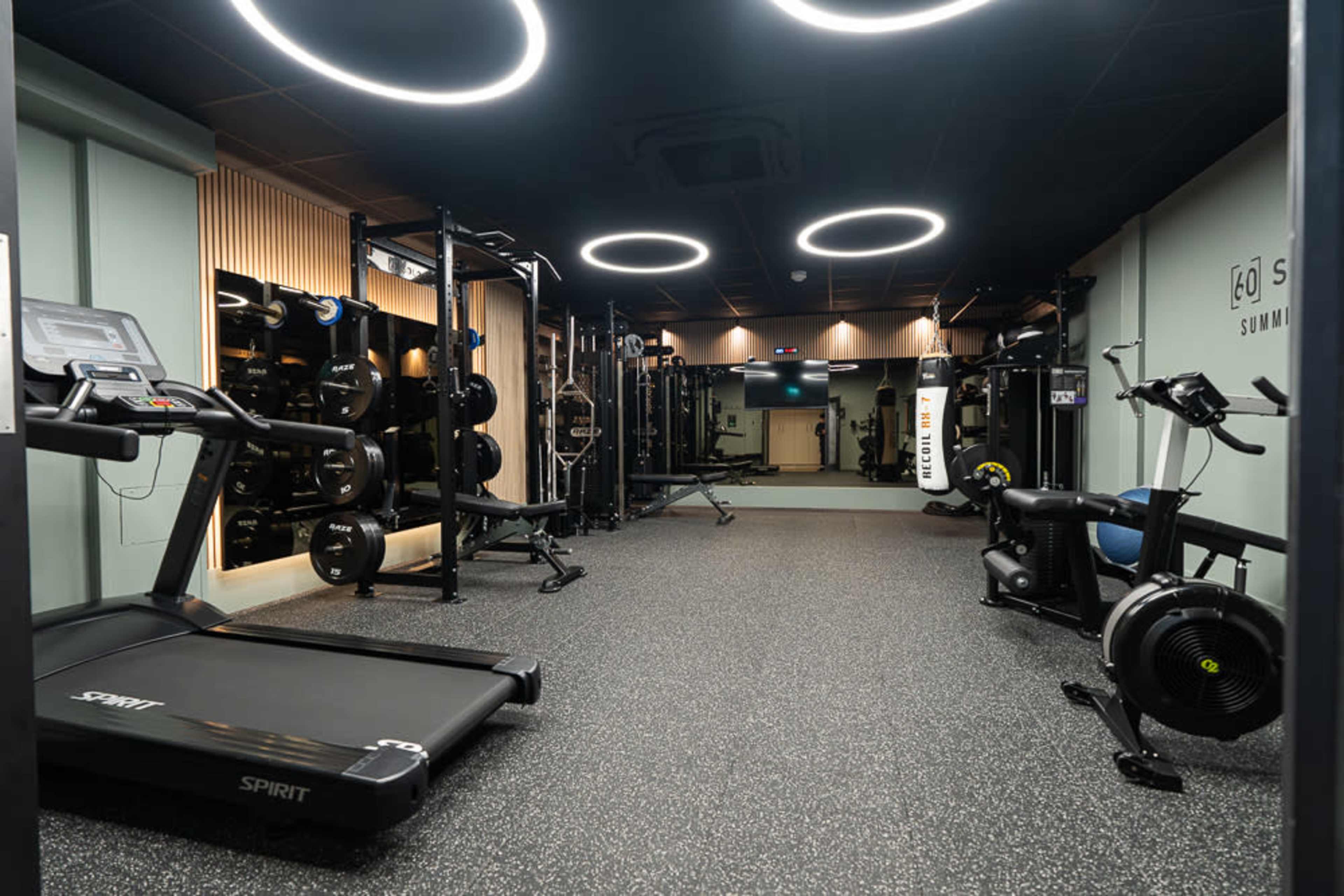 The image shows a modern gym interior equipped with various fitness machines, including a treadmill, weight rack, and rowing machine, surrounded by sleek walls and lighting.