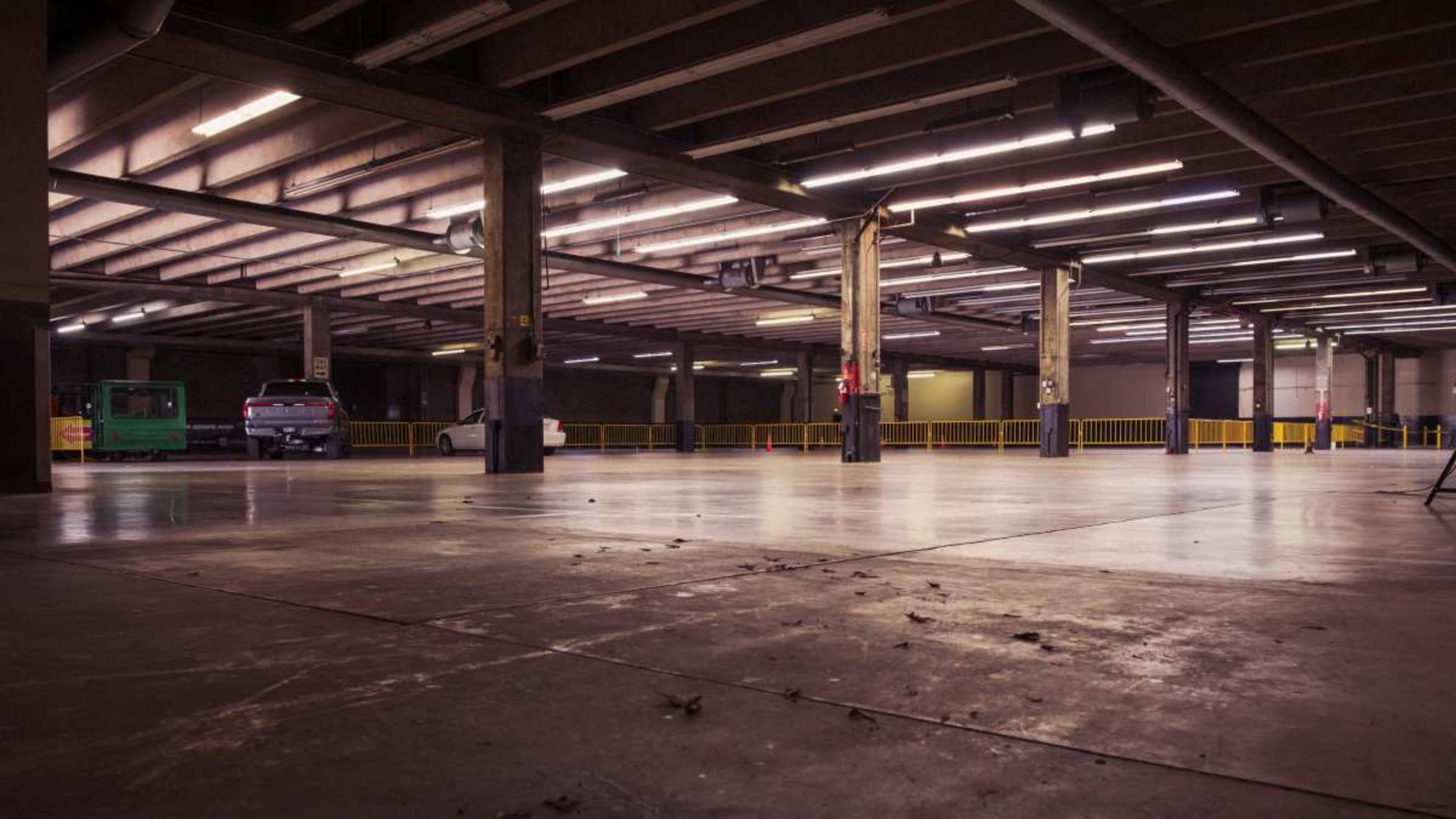 The image shows an empty parking garage with concrete floors, metal pillars, and overhead fluorescent lights.