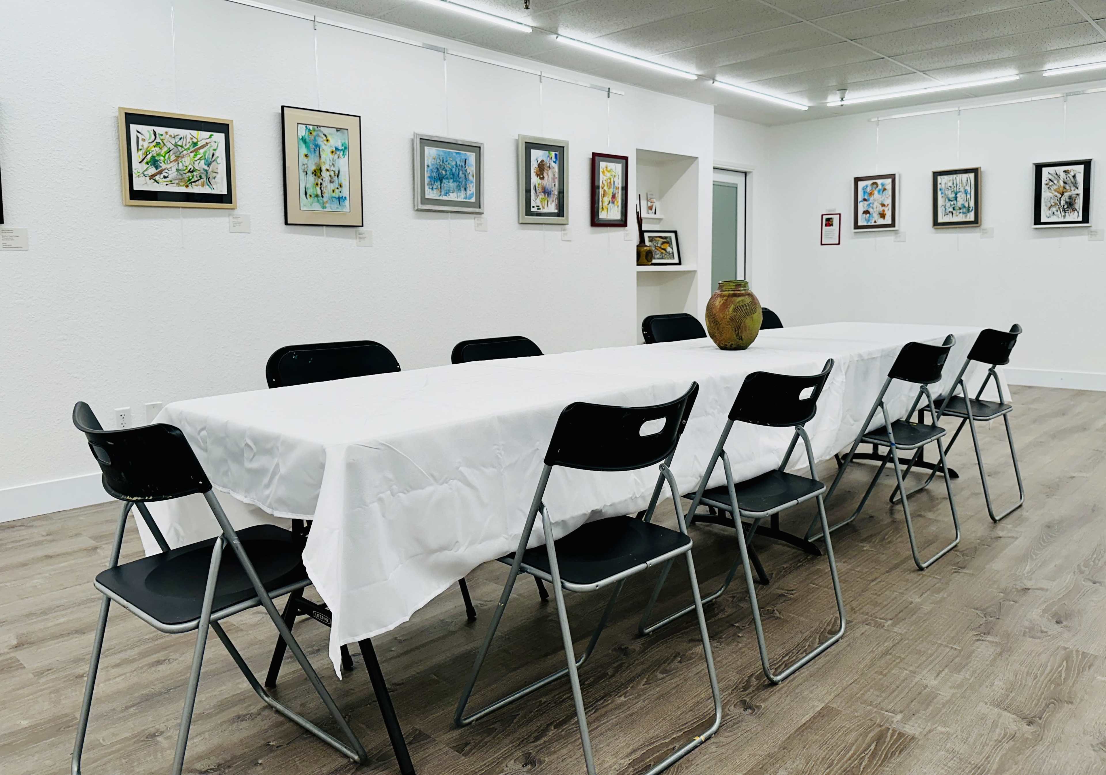 A spacious art gallery with white walls, featuring a long table covered with a white tablecloth and surrounded by black folding chairs, while various framed artworks are hung on the walls.