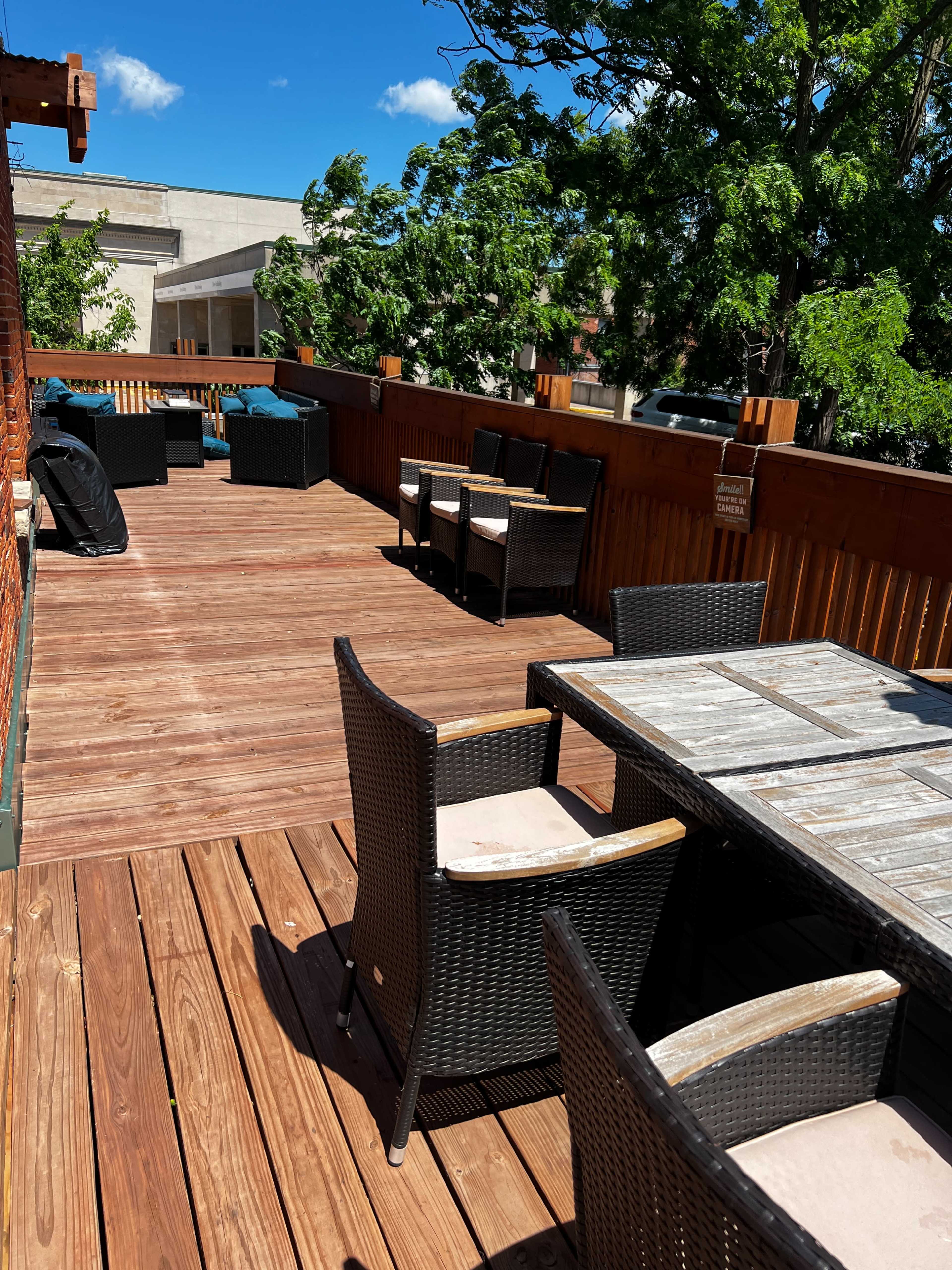 The image shows a wooden deck area with several tables and chairs, surrounded by greenery and blue skies.