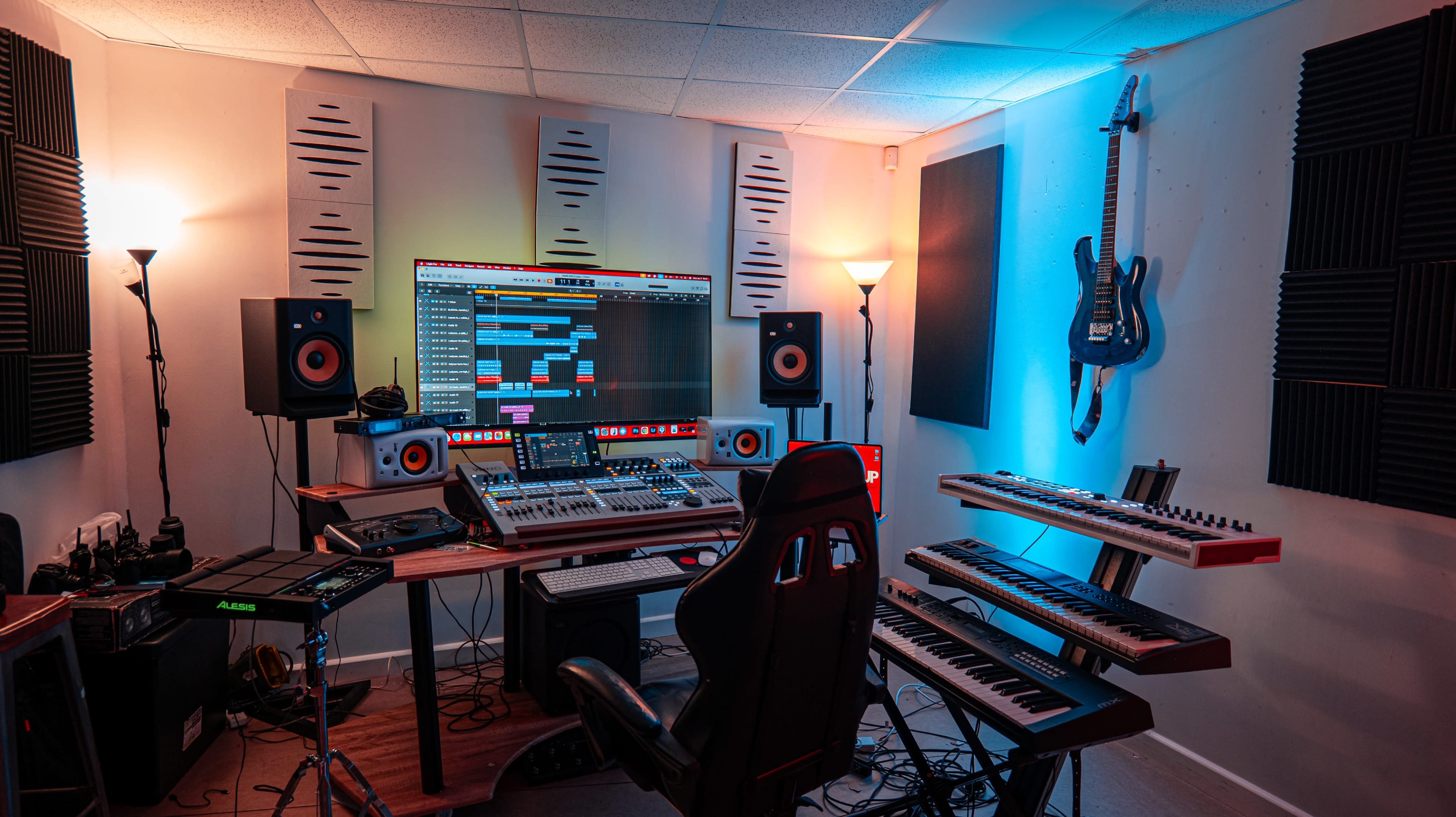 The image shows a music production studio equipped with a large screen, audio monitors, a mixing console, and multiple keyboards, illuminated by colored lights.