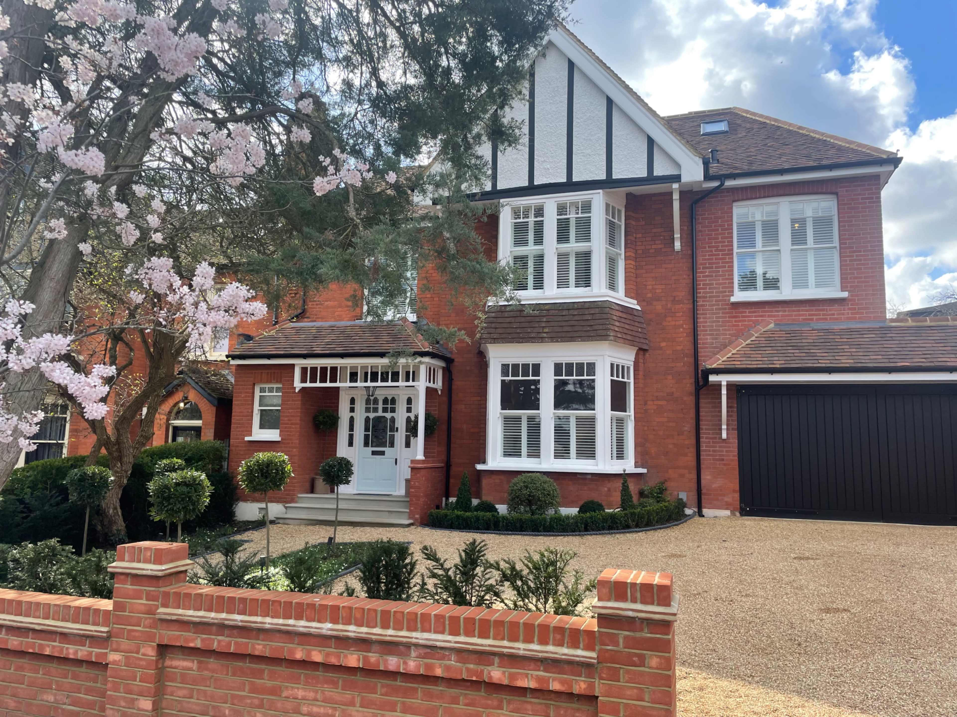 A red-brick house with a gabled roof, large windows, and a neatly landscaped front yard featuring a flowering tree and a gravel driveway.