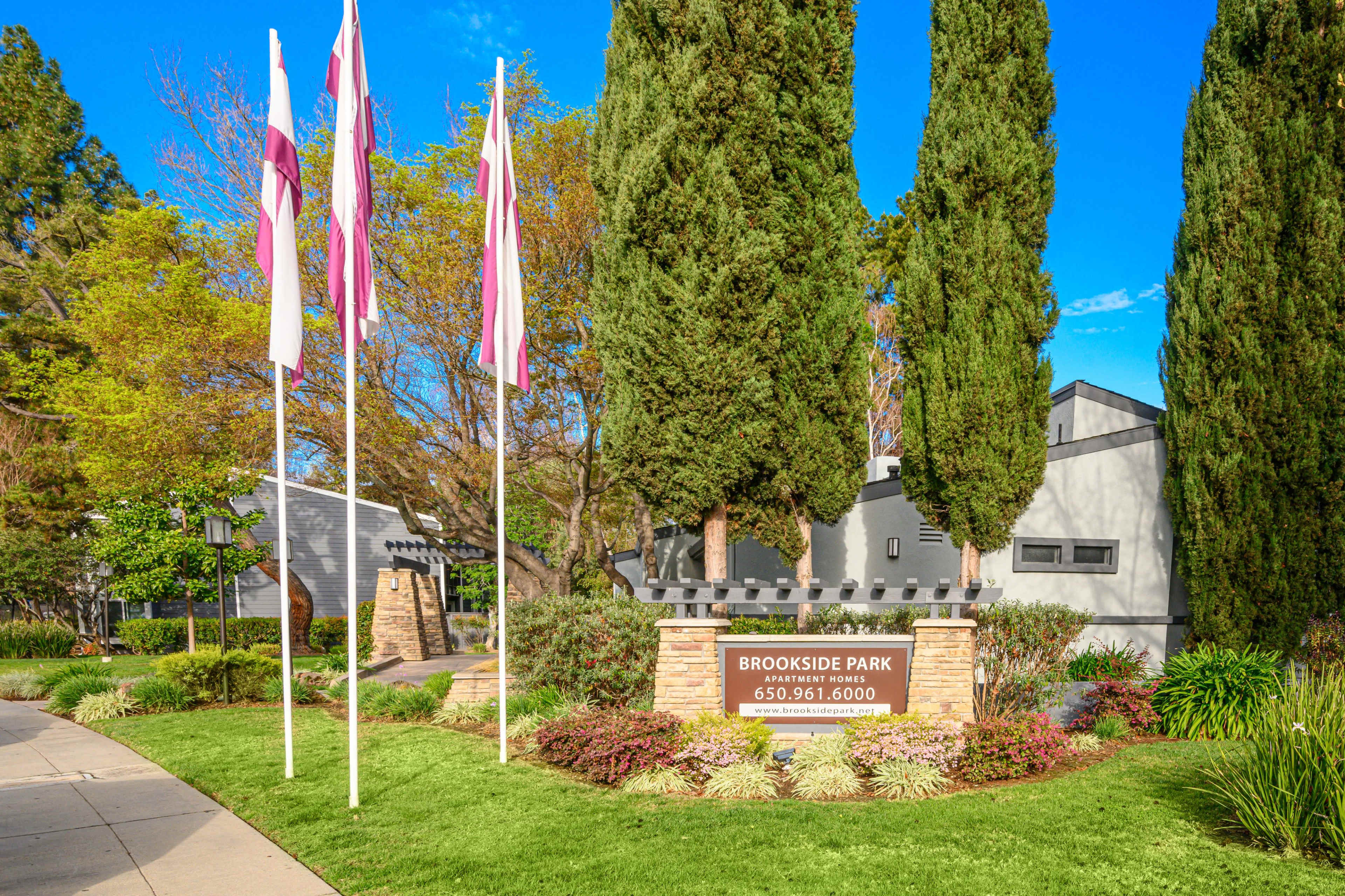 The image shows a landscaped entrance to Brookside Park, featuring tall evergreen trees, decorative flags, and a sign displaying the name and contact number.