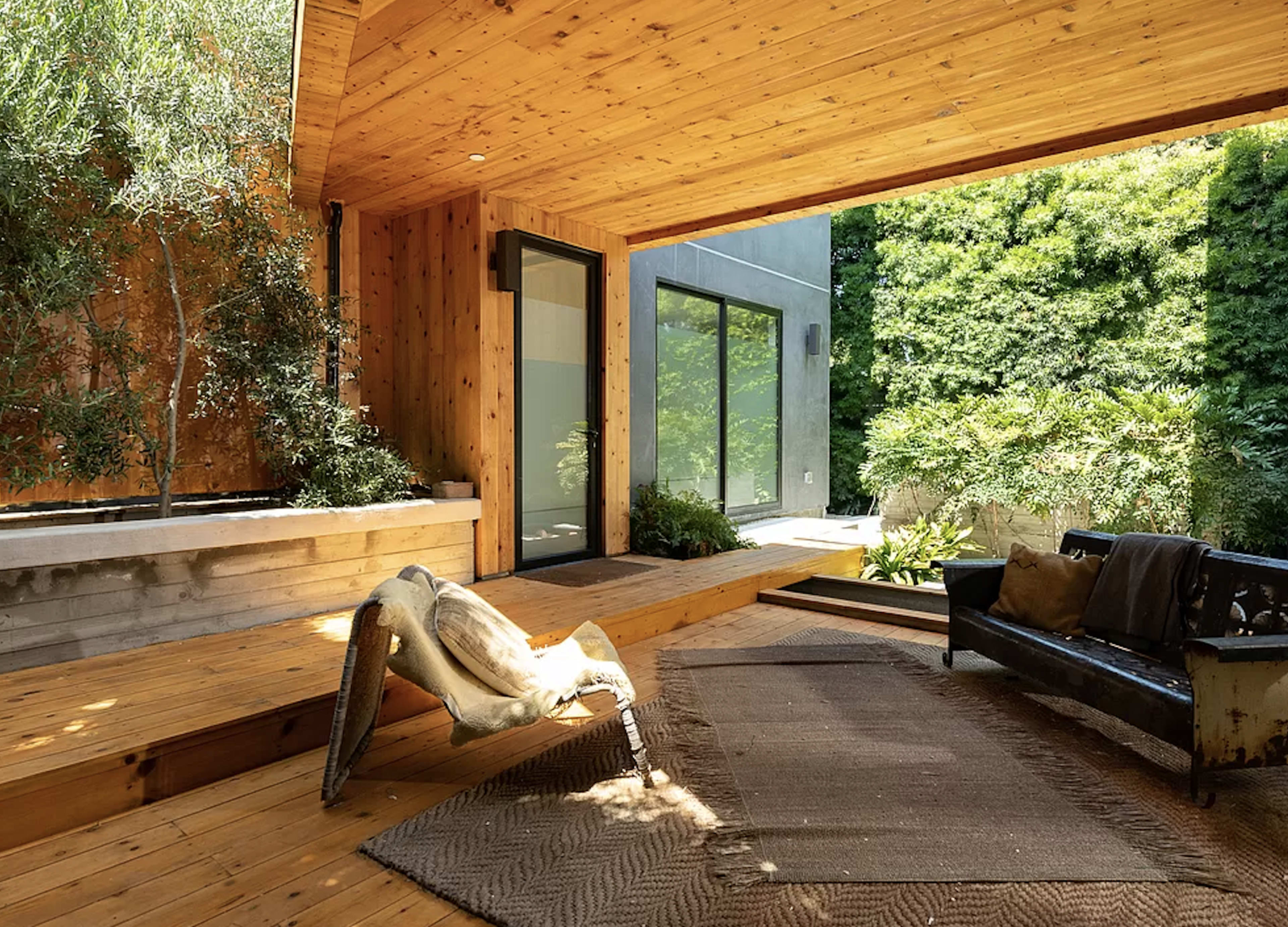 A modern outdoor living space with wooden decking, a sleek black sofa, and lush greenery in the background.