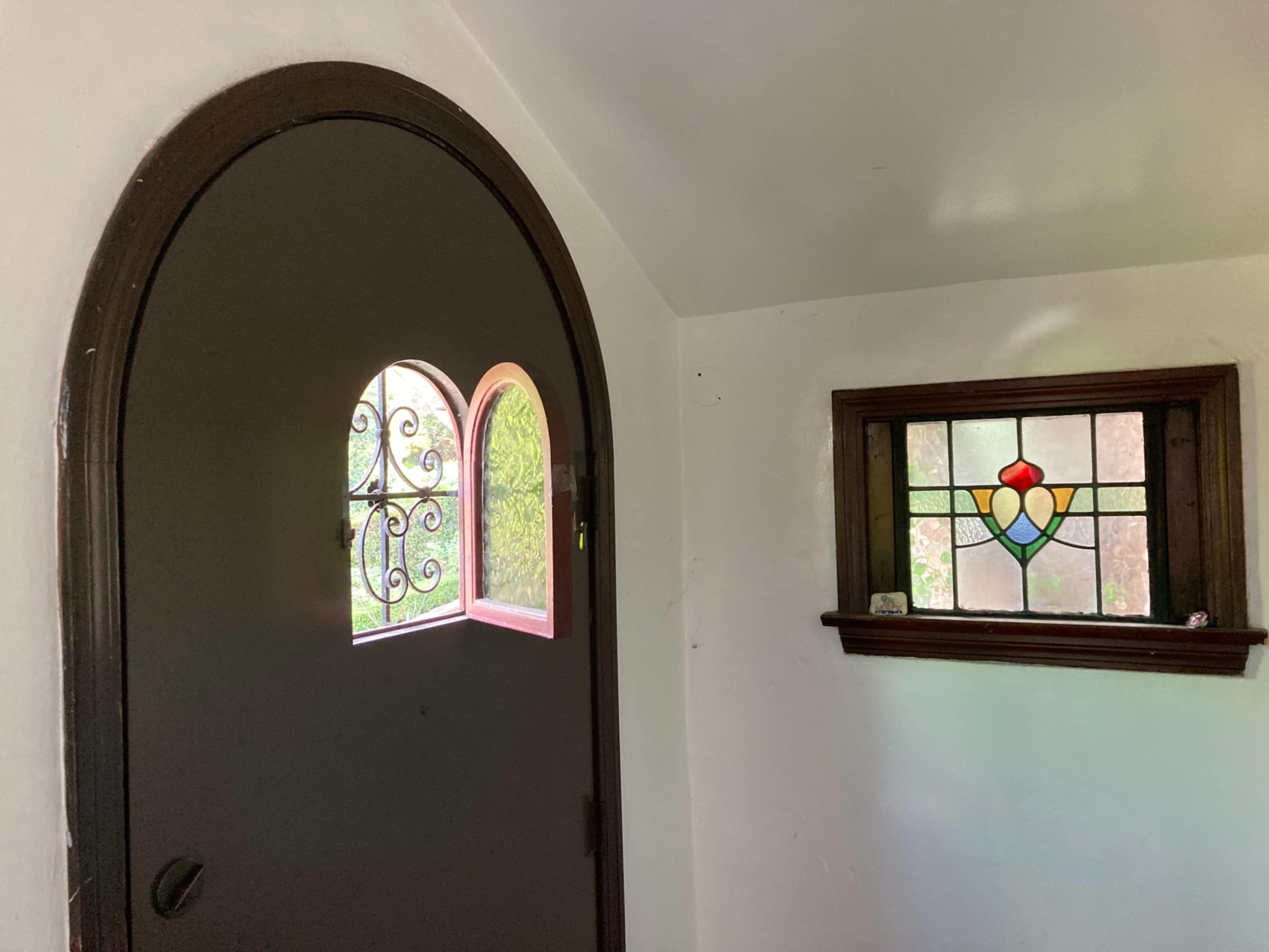 An arched wooden door with decorative windows and a stained glass window set into a white wall.