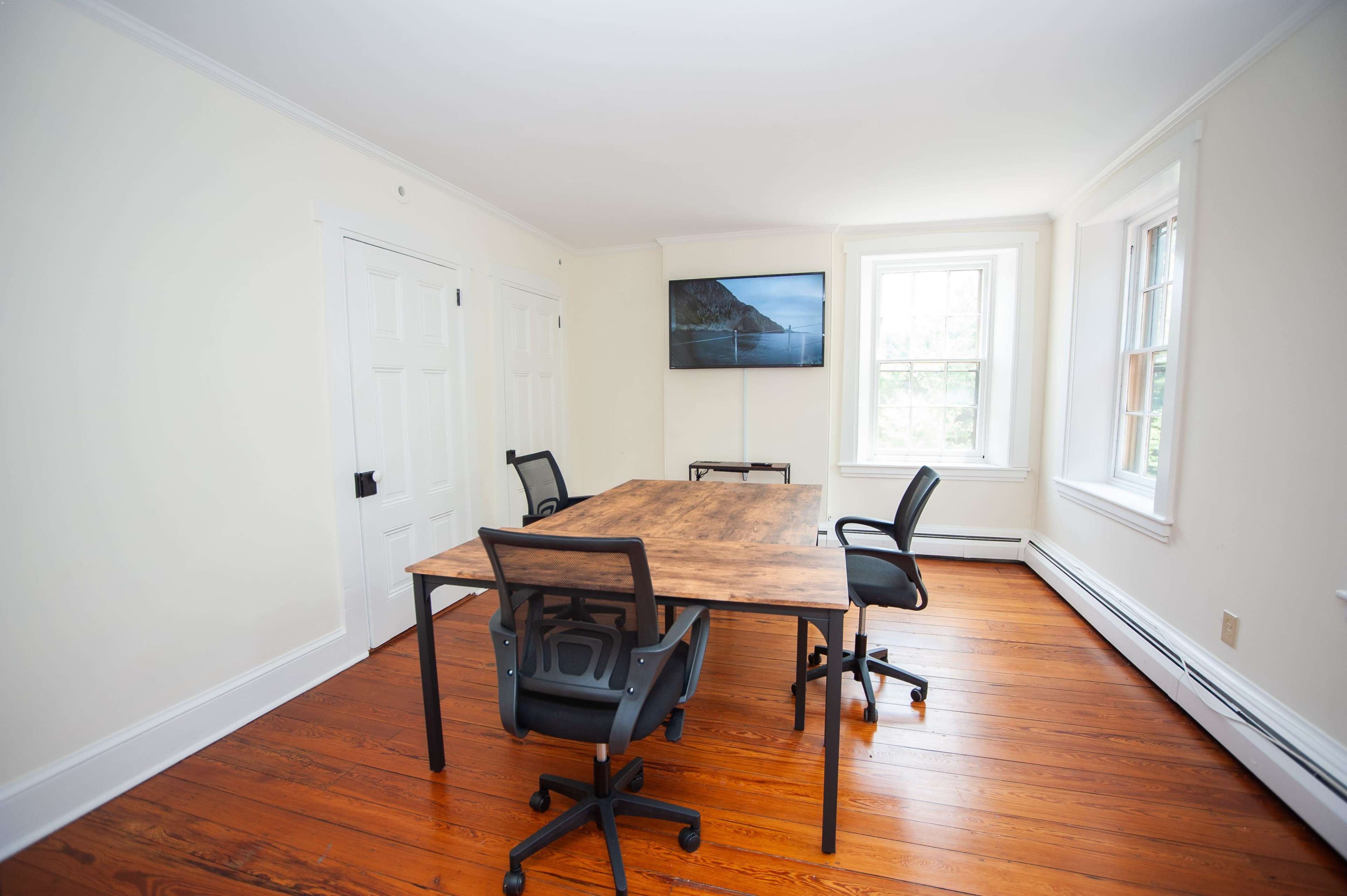 A simple conference room with a wooden table and four black chairs, a television mounted on the wall, and windows allowing natural light.