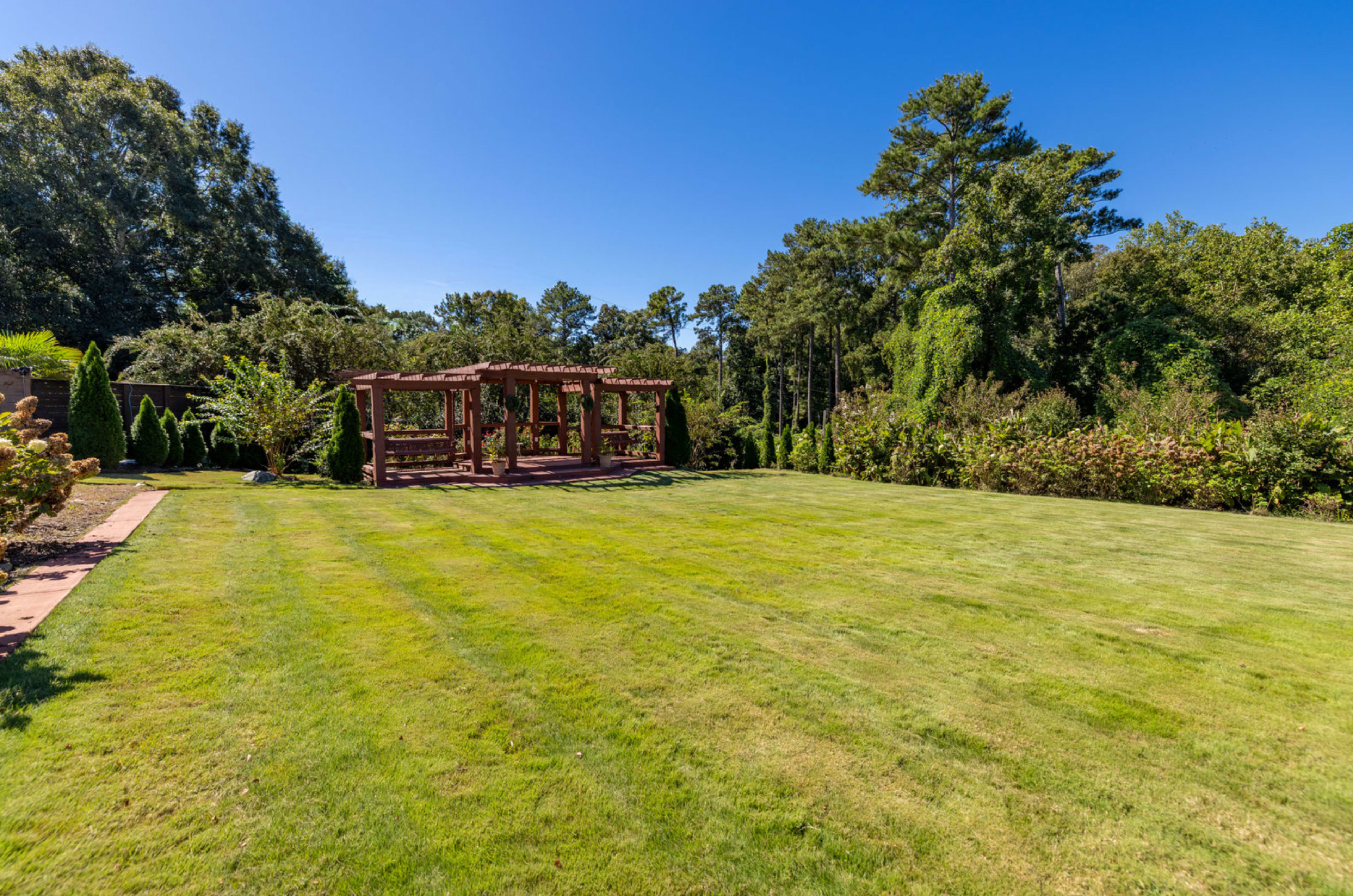 The image shows a well-maintained lawn with a wooden pergola in the background, surrounded by lush greenery and trees under a clear blue sky.