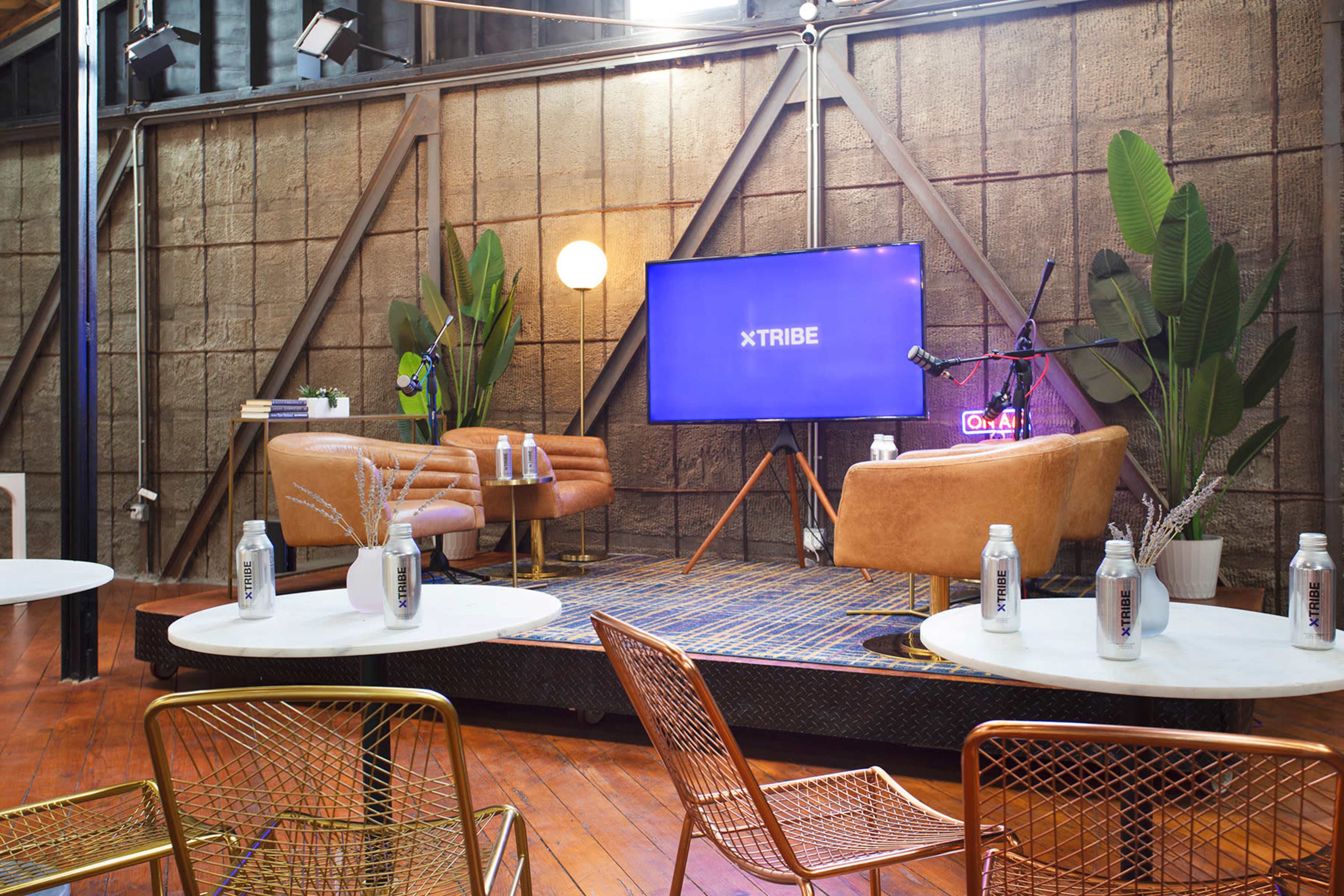 A modern lounge area features two brown sofas facing a television screen, surrounded by potted plants and small white tables with metallic chairs.