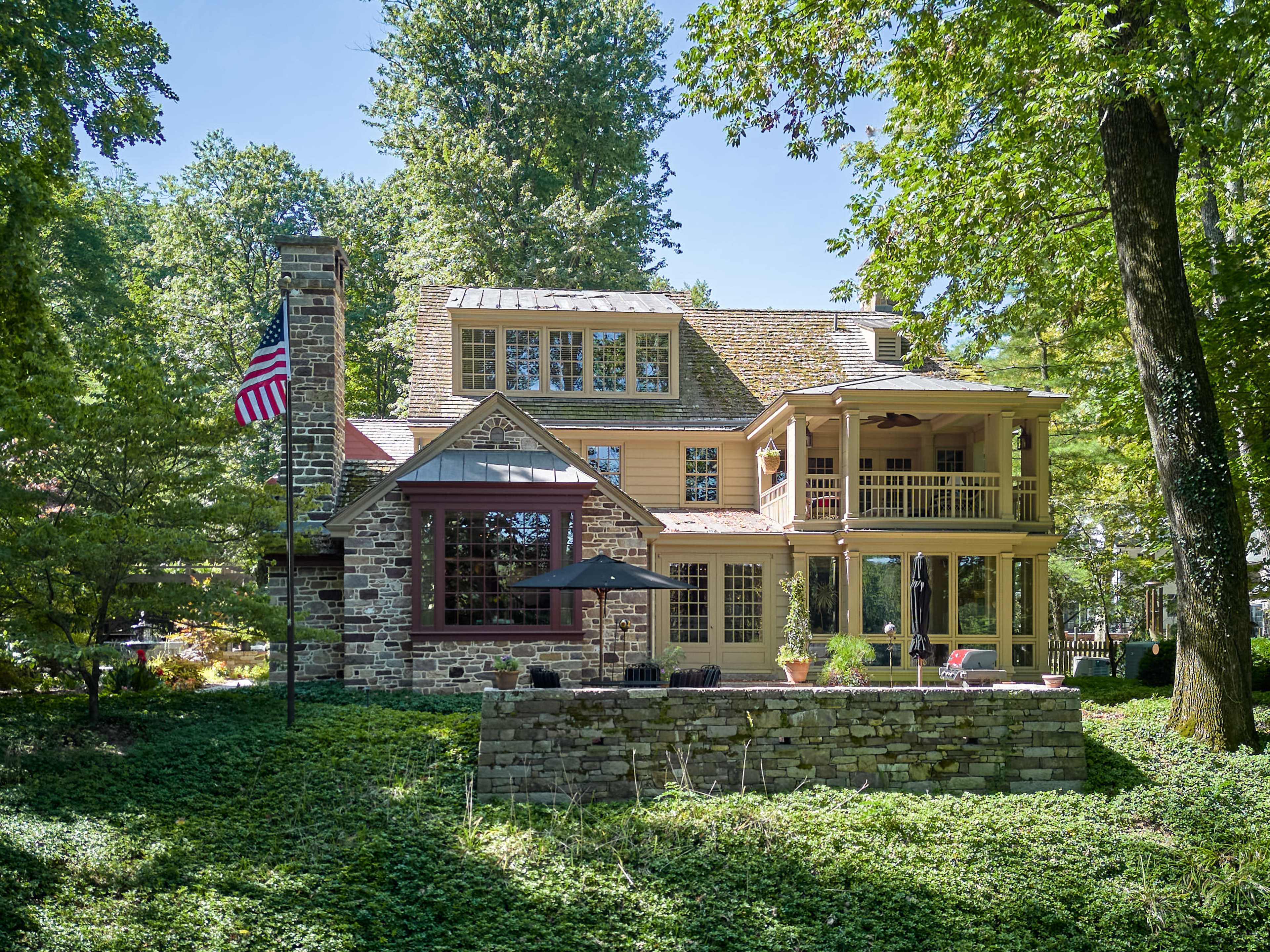 A large, multi-story stone and wood house with a porch, surrounded by trees and greenery.