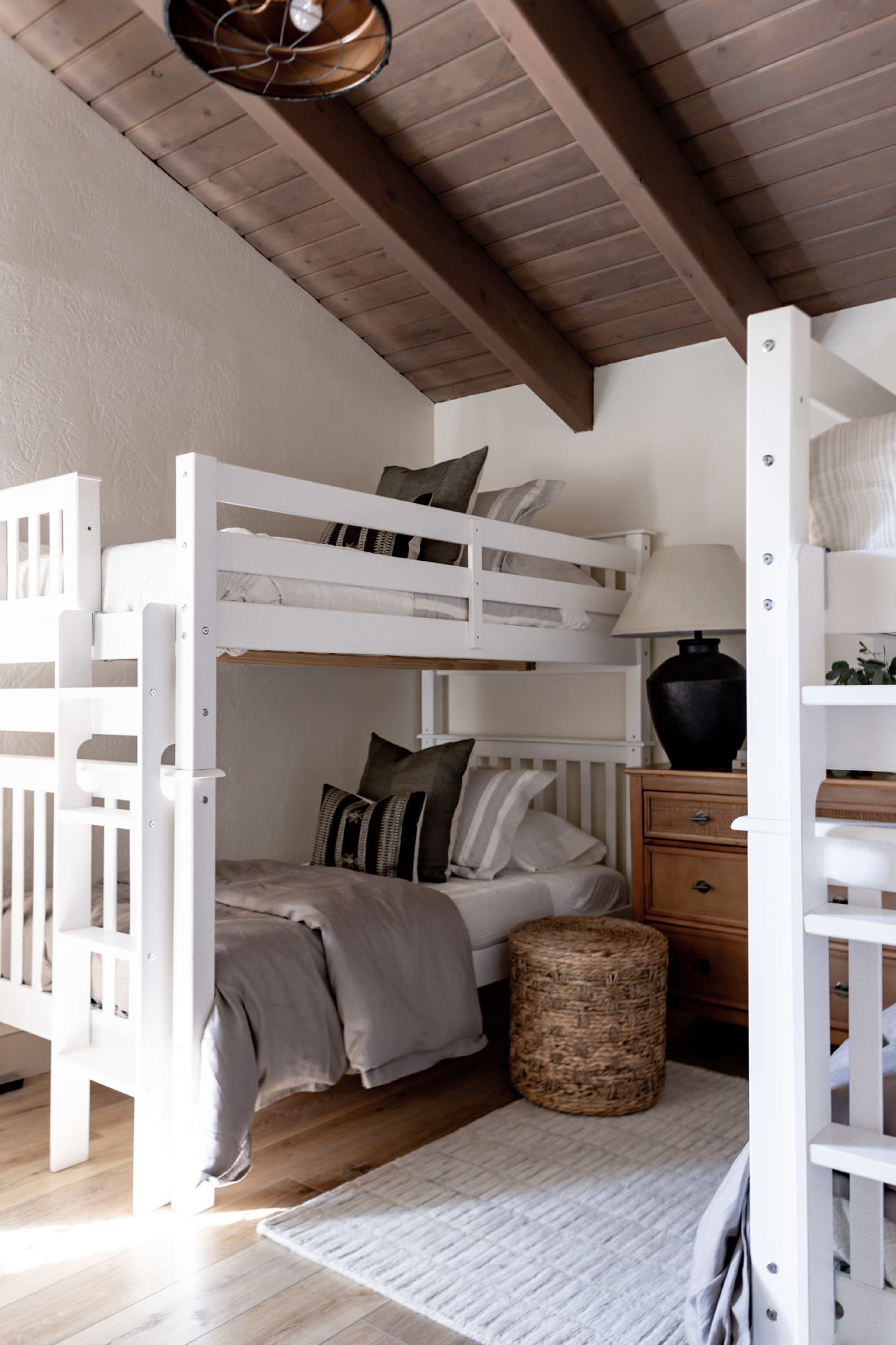 The image shows a cozy bedroom with a lofted bunk bed, a single bed beneath, a wooden dresser, and a woven stool.