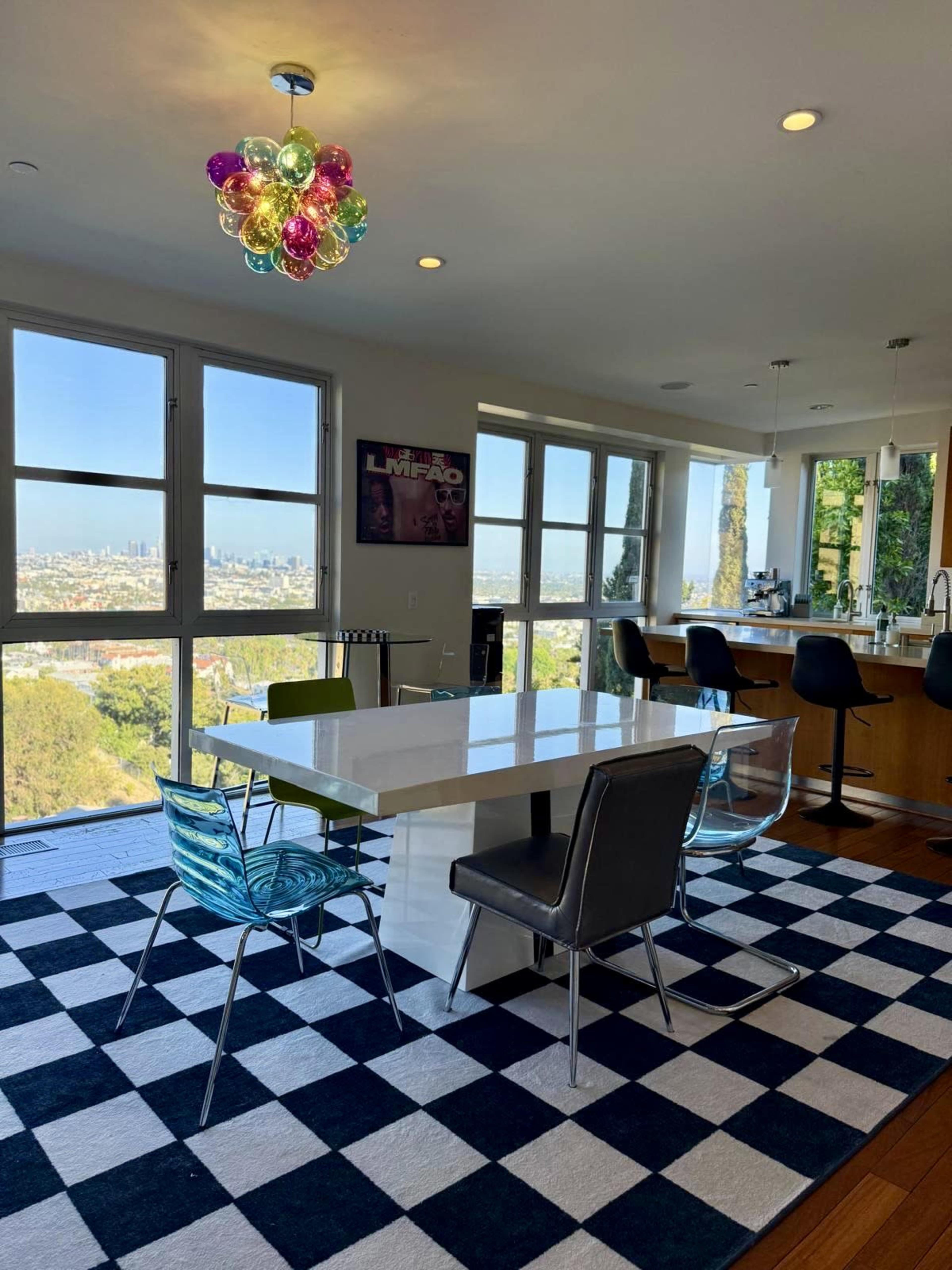 The image shows a modern dining area with a white table, colorful chairs, and a large window providing a view of a cityscape.