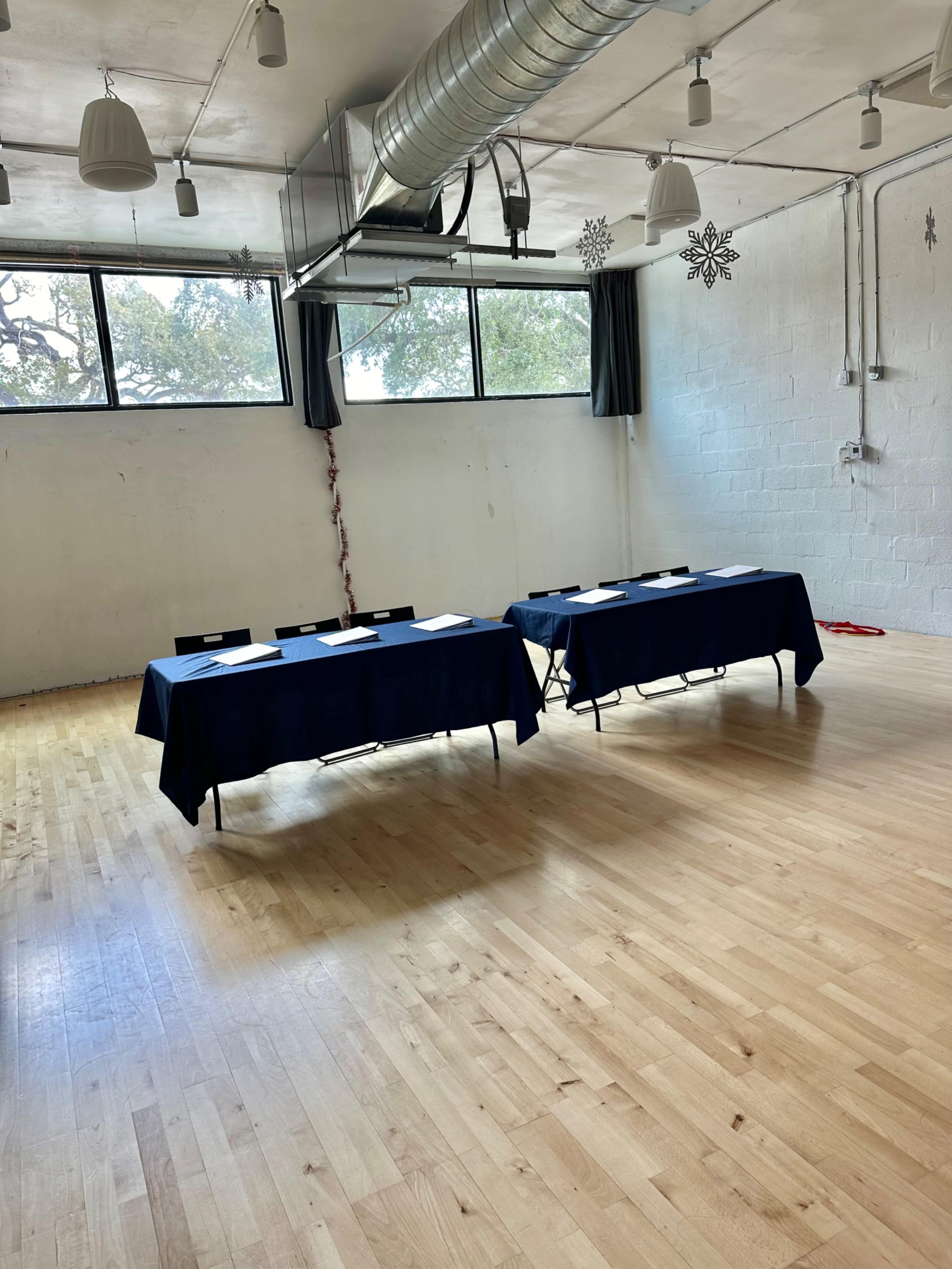 The image shows a spacious room with a wooden floor, featuring two tables draped with blue tablecloths, placed parallel to each other under a row of windows.