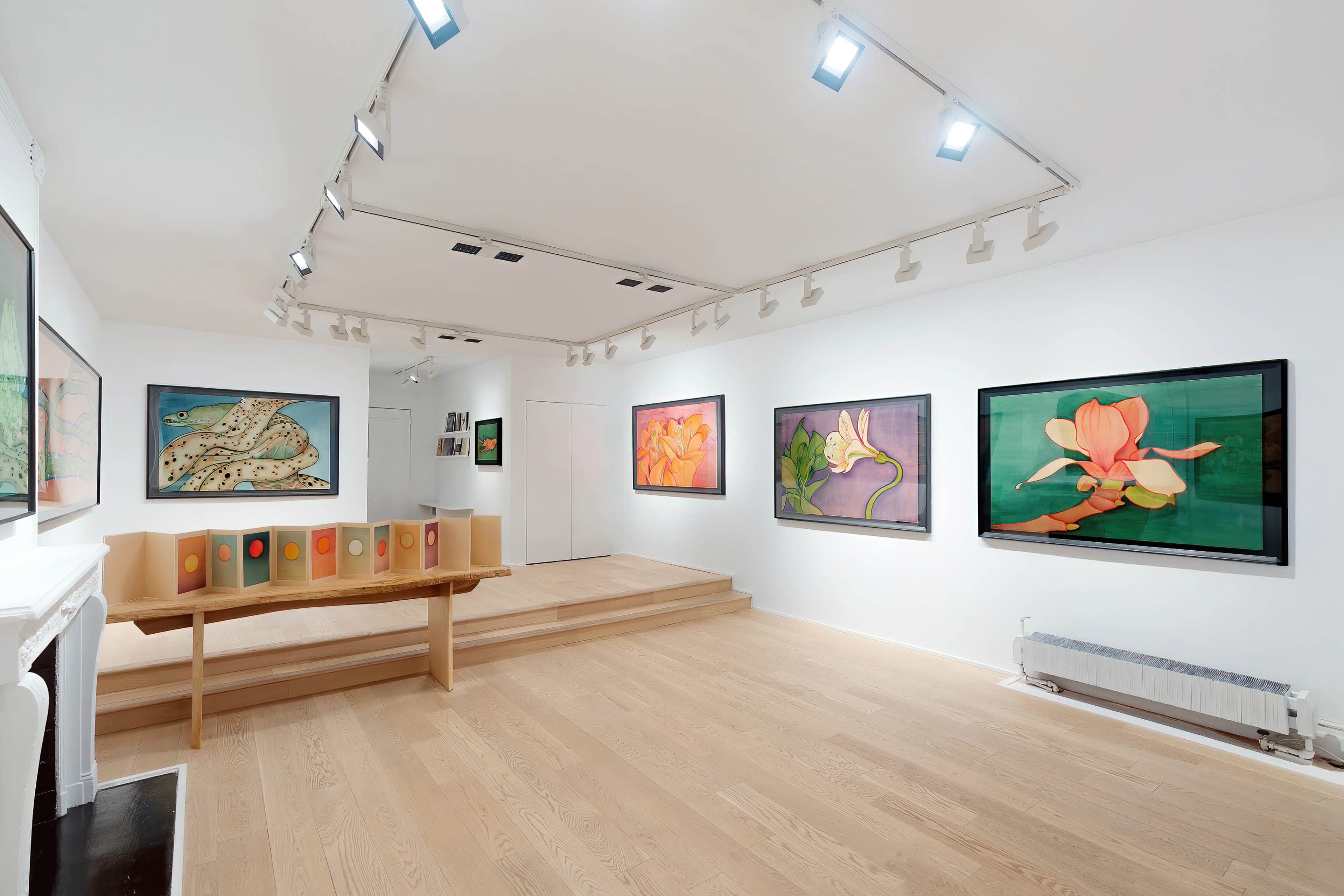 An art gallery with brightly lit walls displaying framed floral paintings and a wooden table featuring displayed art pieces.